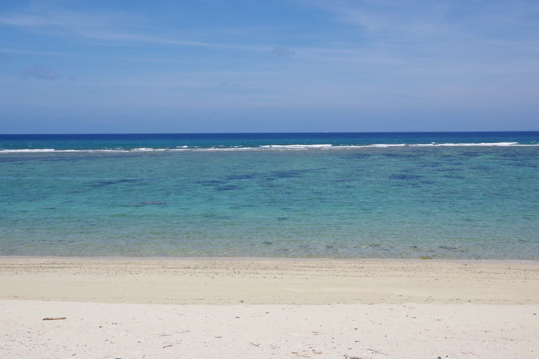 White sand gives way to bright shallows and a band of reef off the coast at Ogimi, with deep blue open sea beyond under a clear sky