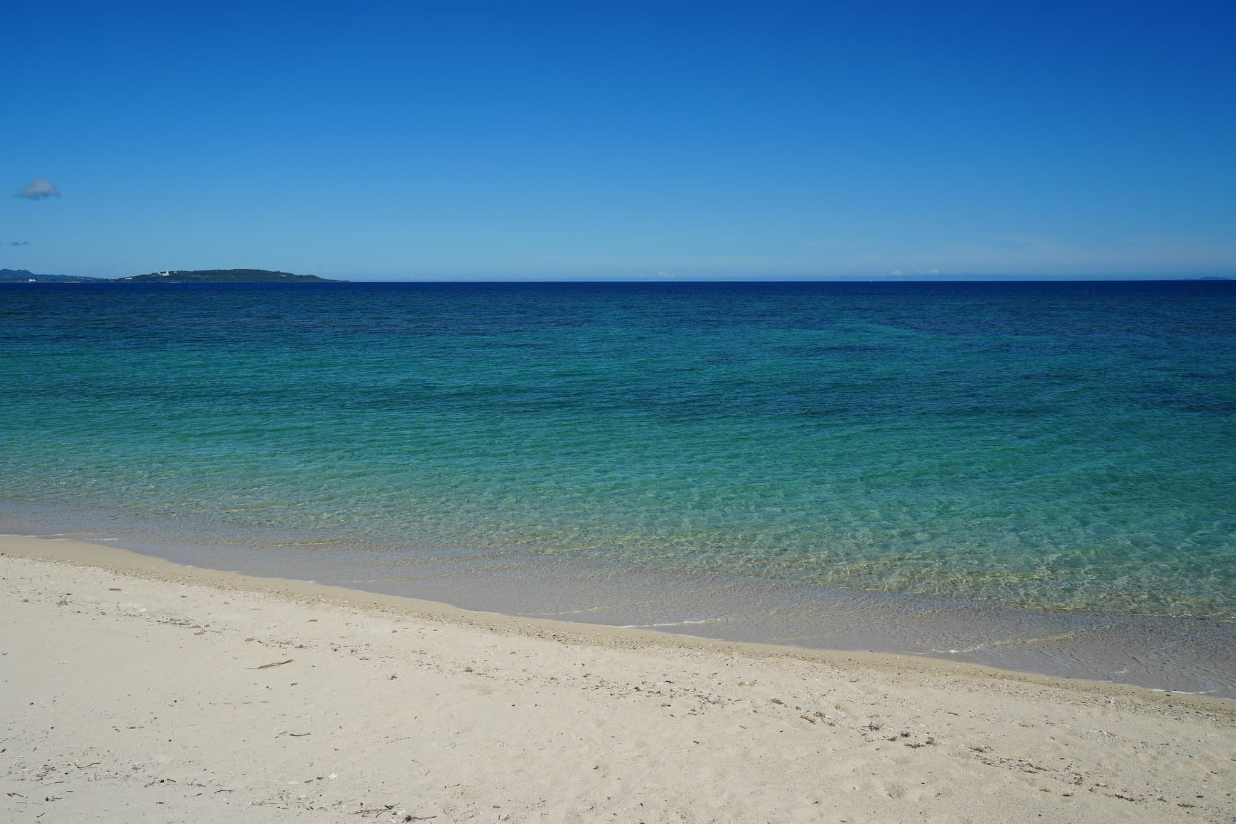 Clear turquoise water laps a quiet white beach in Ogimi on a summer day, and low islands sit on the far horizon