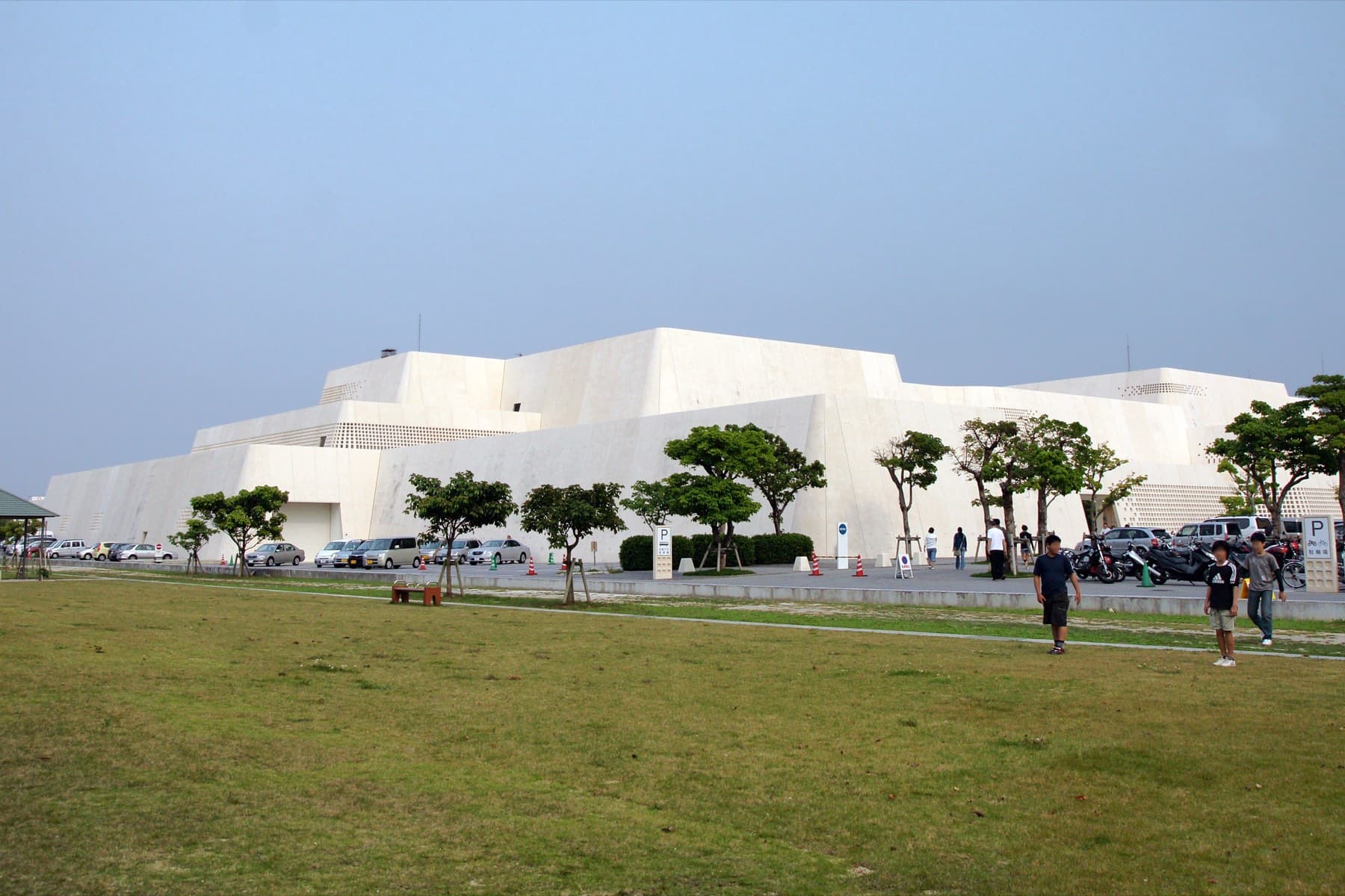 The white stepped bulk of the Okinawa Prefectural Museum and Art Museum rises like a gusuku wall beyond a wide lawn and young trees in Omoromachi