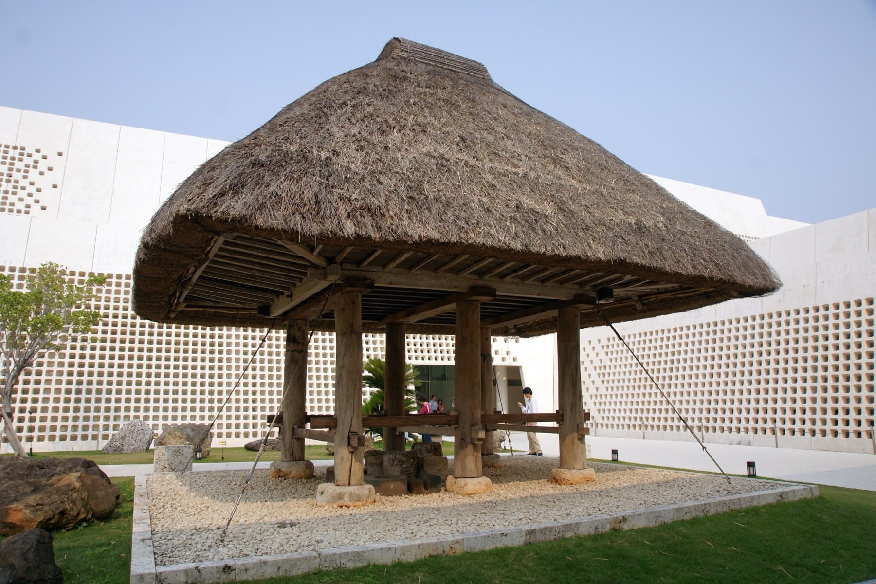 A thatched takakura granary on wooden stilts stands on a gravel garden in the courtyard of the Okinawa Prefectural Museum, against its white screen walls