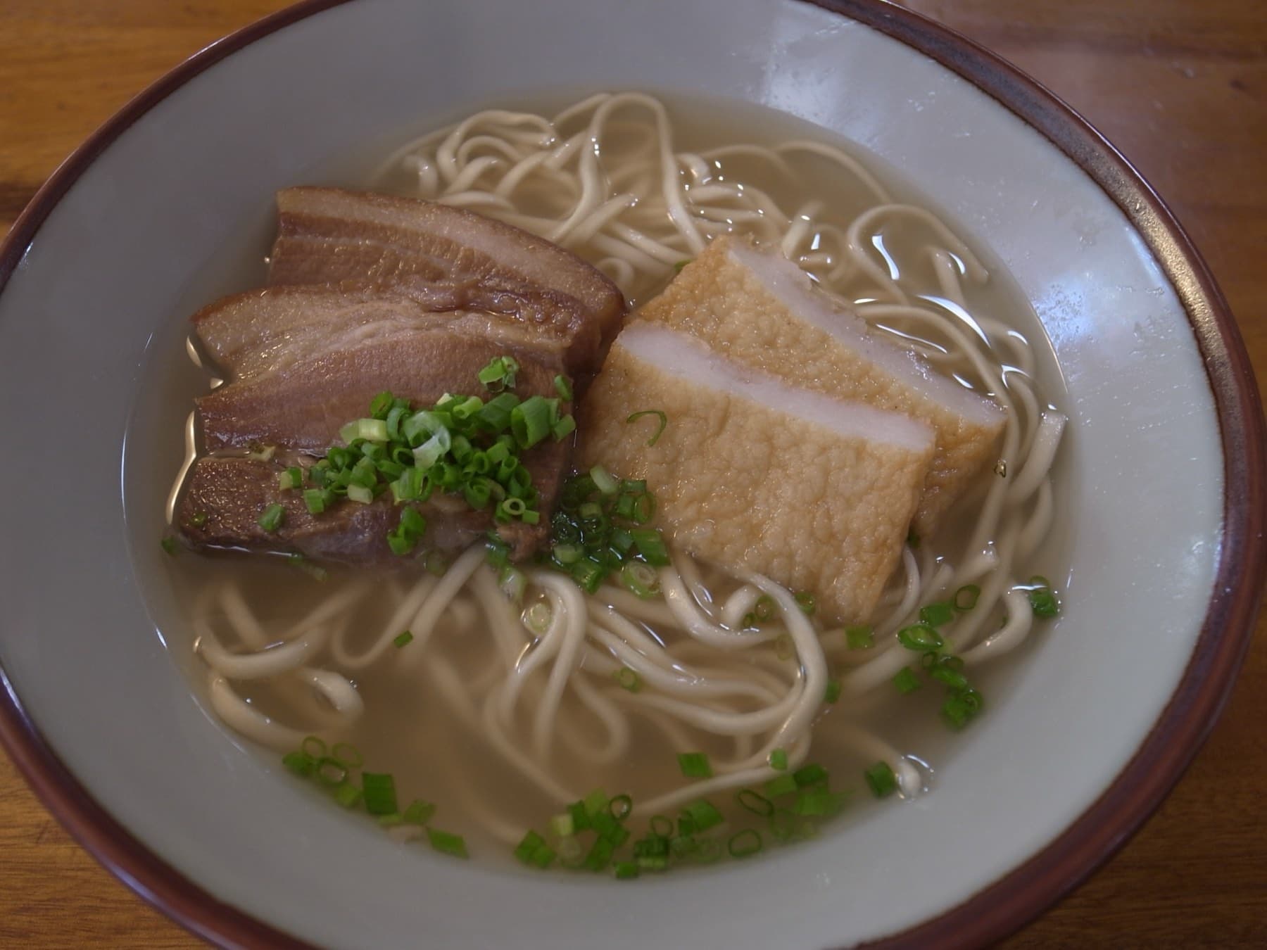 Two slices of braised pork belly and a block of fried tofu rest on thick wheat noodles in a pale broth scattered with chopped green onion