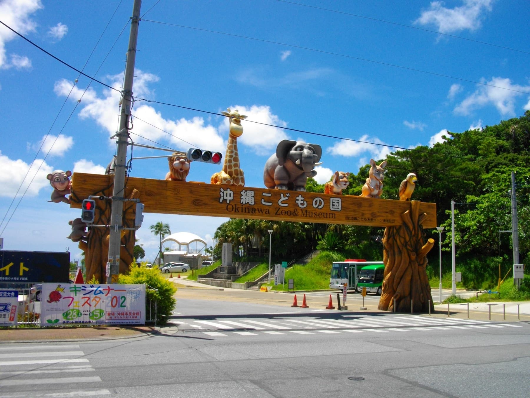 A carved wooden arch topped with giraffe, elephant and monkey figures spans the road at the entrance to the Okinawa Zoo and Museum under a summer sky