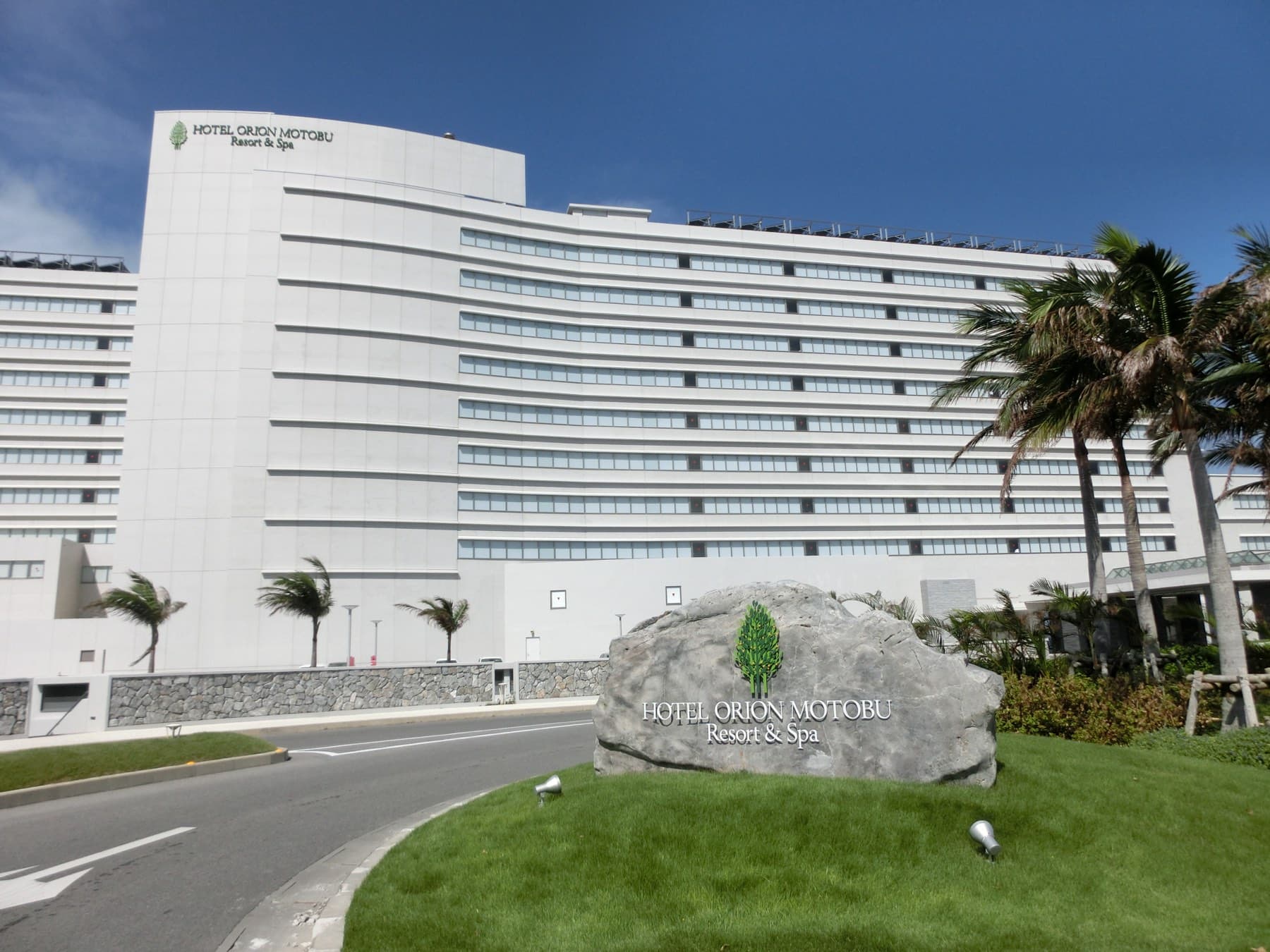 The long white curve of Hotel Orion Motobu Resort & Spa rises behind palms and a stone sign at its entrance beside Ocean Expo Park