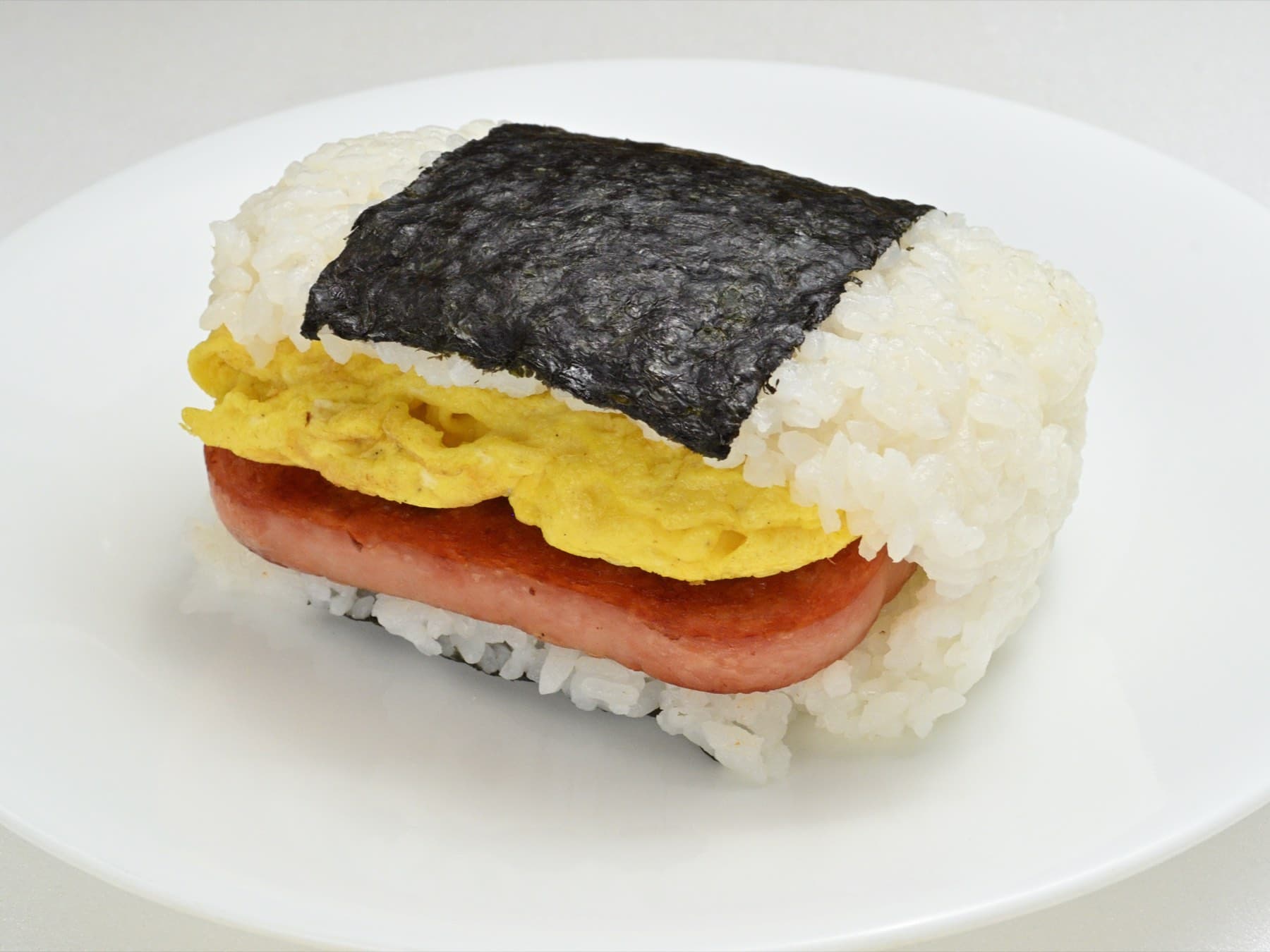 A thick slice of luncheon meat and a folded omelette are pressed between two layers of white rice and bound with a strip of nori on a white plate