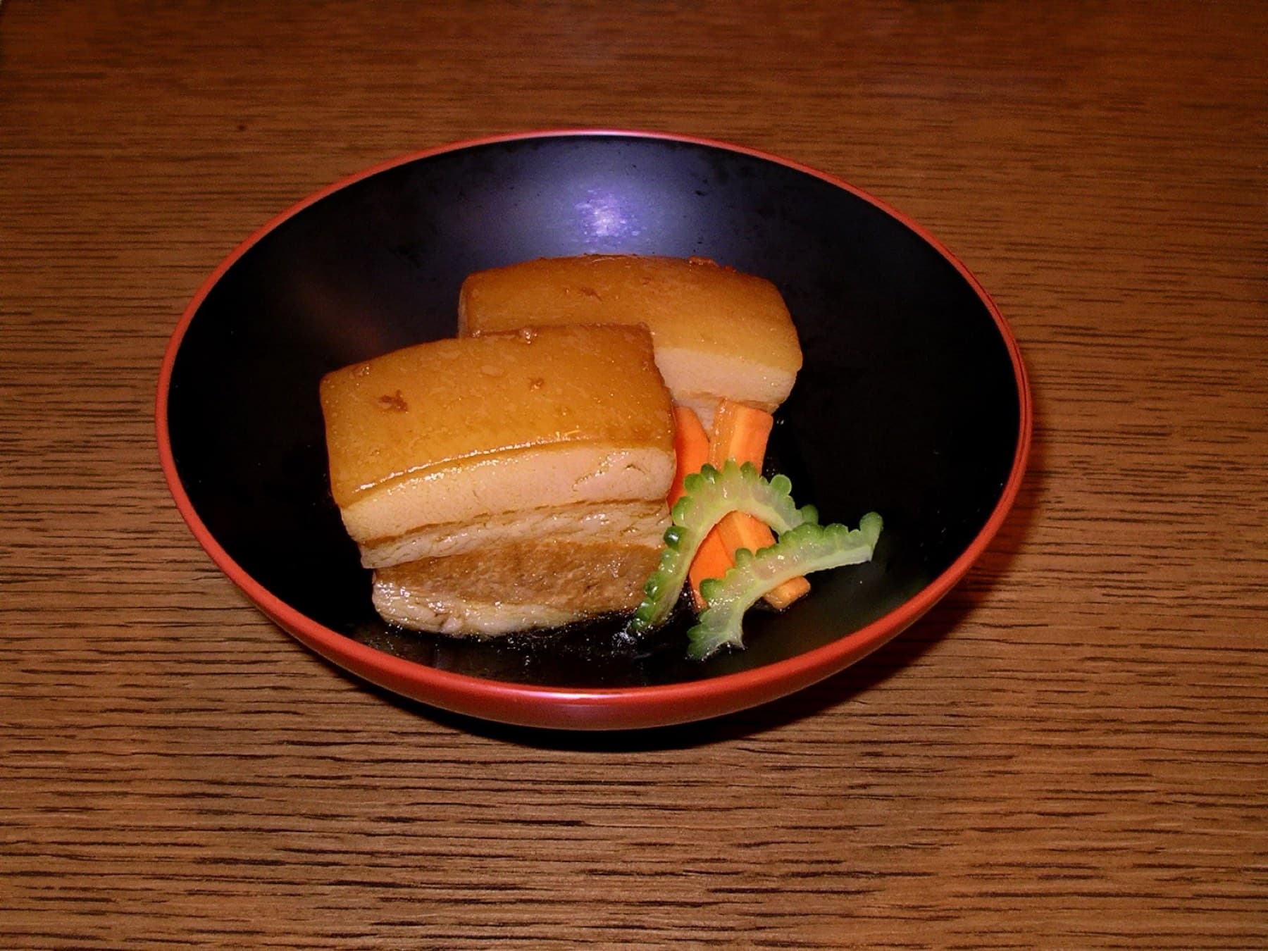 Two glazed cubes of slow-braised pork belly sit with carrot and a slice of bitter melon in a black and red lacquer bowl on a wooden table