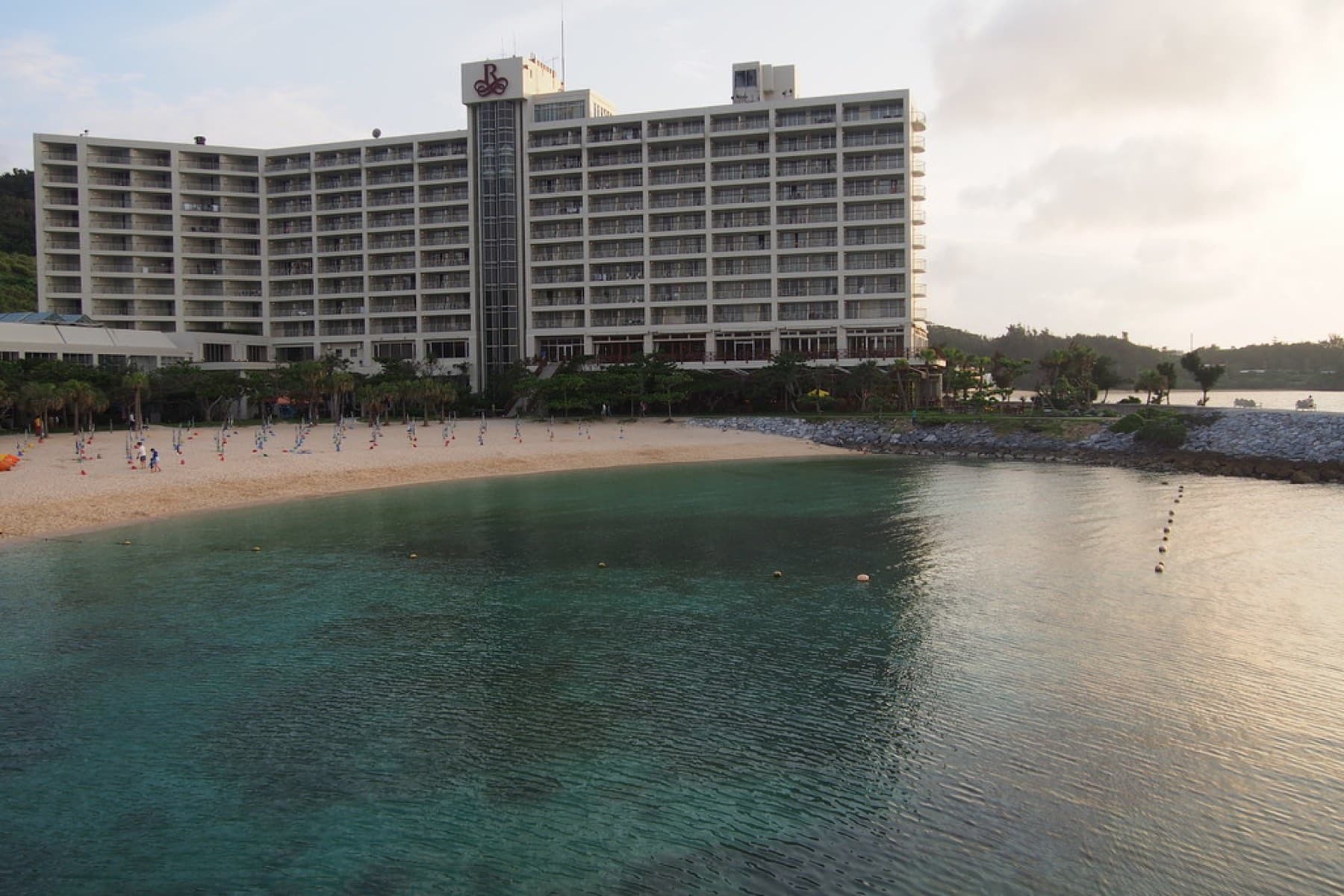 Calm green water fills the sheltered cove in front of the Renaissance Okinawa Resort tower, where sun loungers line a sandy beach in soft evening light