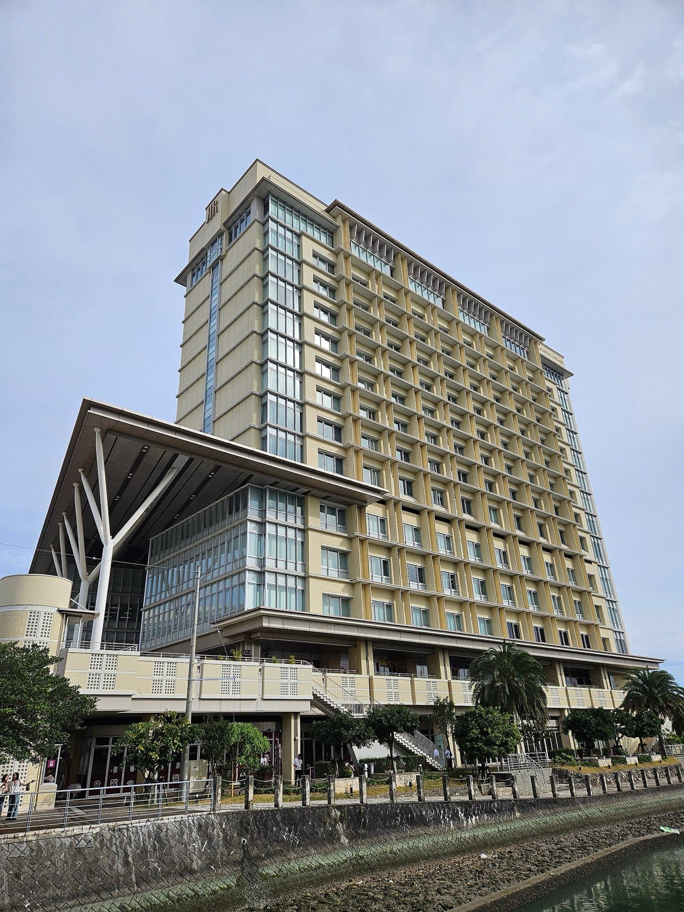 The Rihga Royal Gran Okinawa tower stands above a low glass podium with a sweeping white roof beside the Kumoji River in central Naha