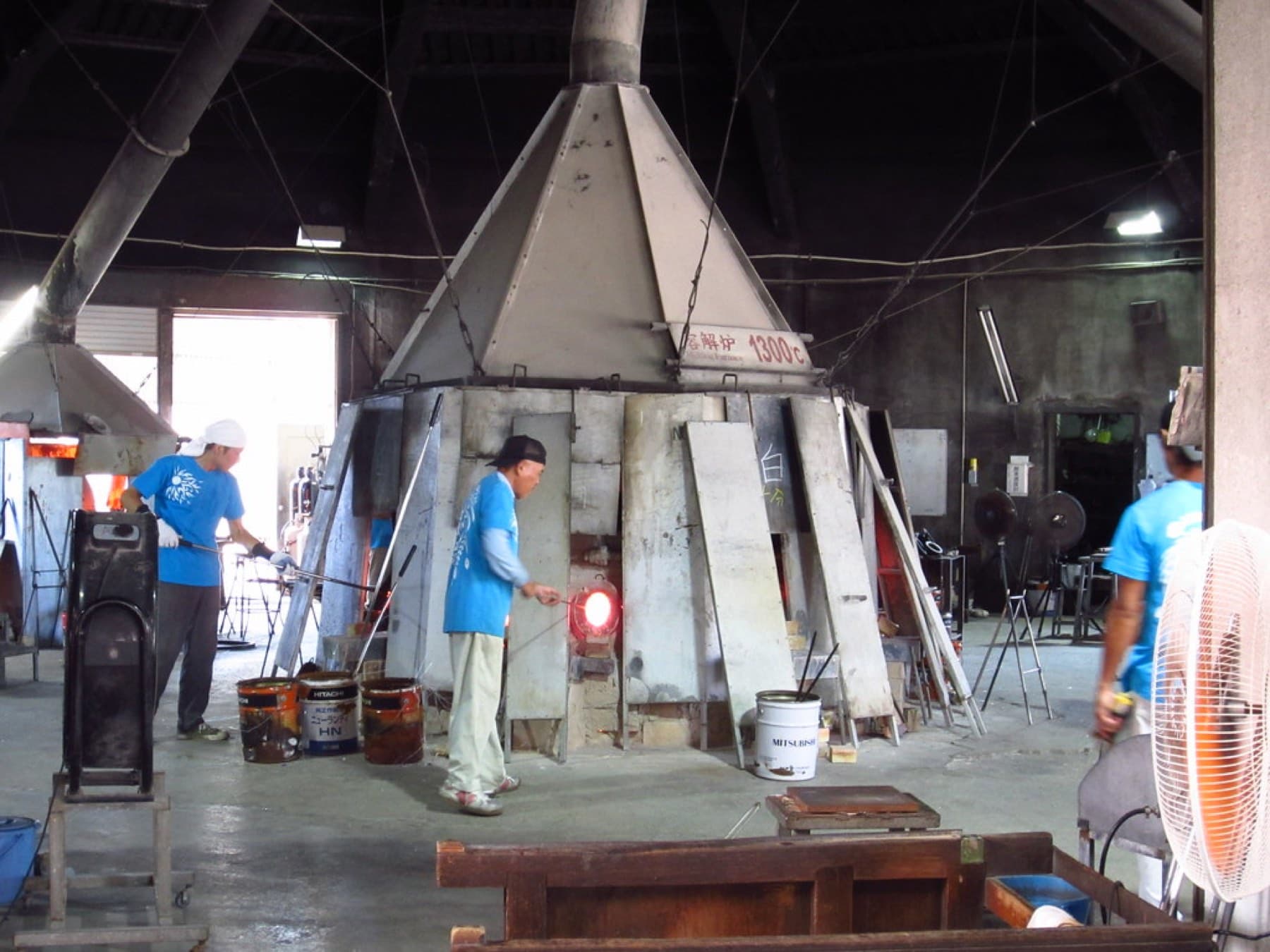 Glassblowers in blue shirts work at the glowing mouths of a large hooded furnace on the concrete floor of a glass workshop in Itoman