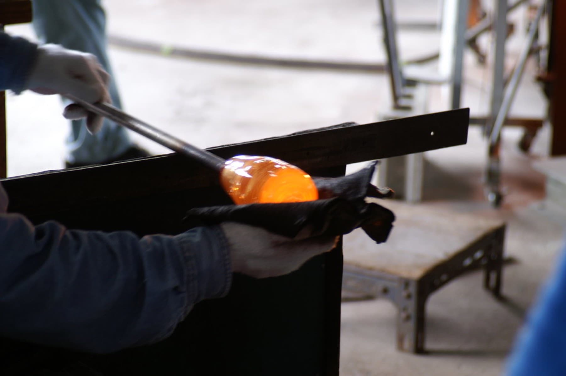 A glassblower shapes a glowing orange gather of molten glass on a steel pipe with a folded wet pad at a workbench in Tamagusuku