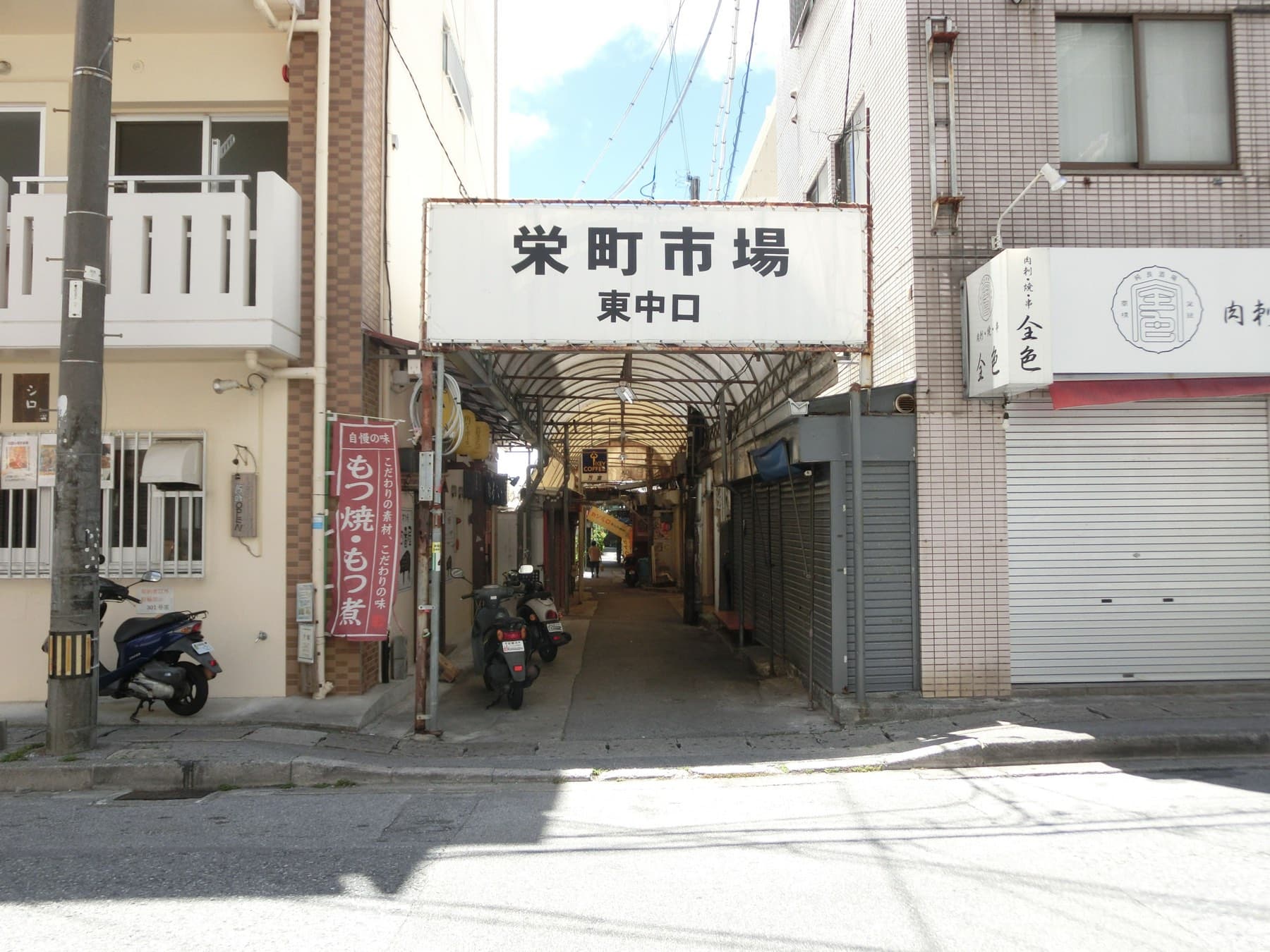 A white sign marks the east entrance of Sakaemachi Market in Naha, where a narrow covered lane runs back between shuttered stalls and parked scooters