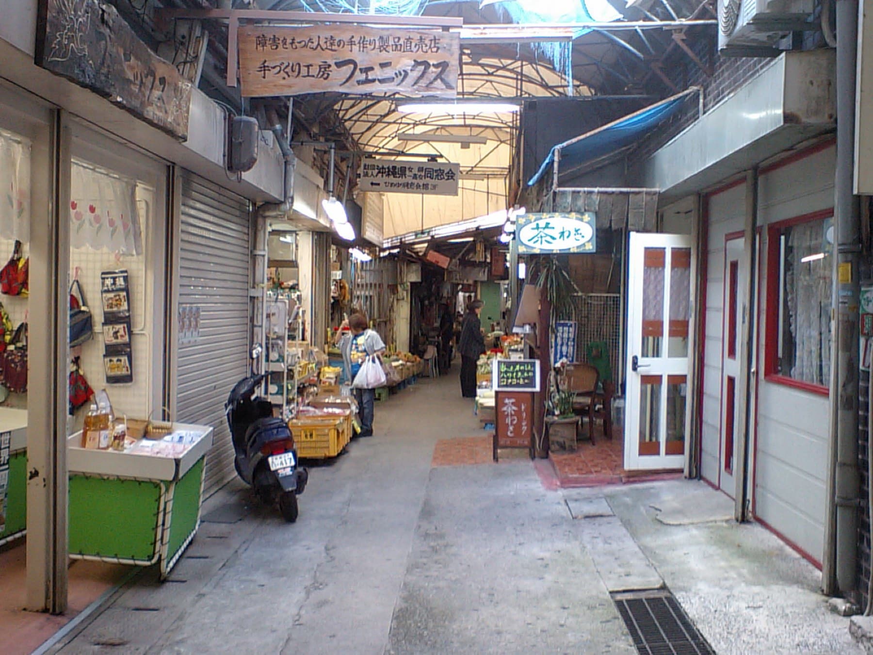 Inside the covered lanes of Sakaemachi Market a shopper walks past produce stands and a parked scooter in the soft light under the arcade roof