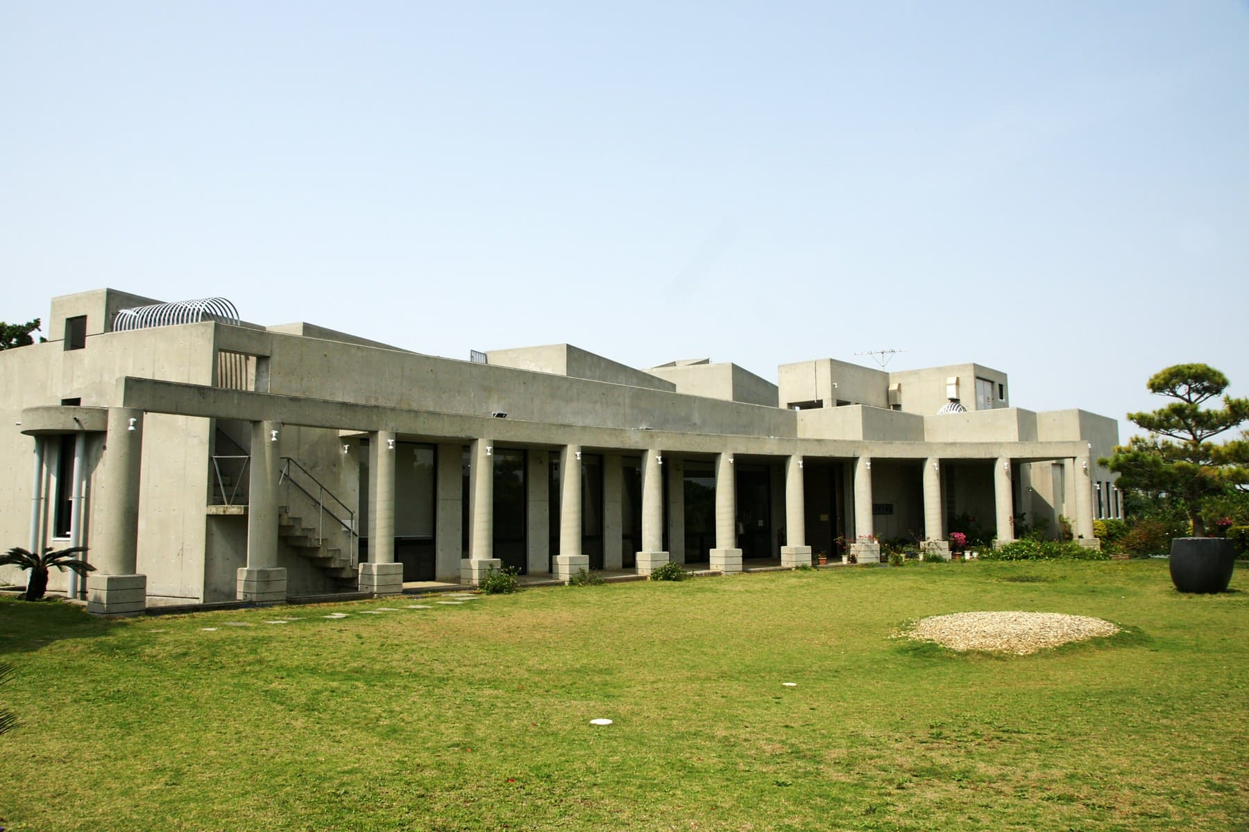 The low grey concrete colonnade of the Sakima Art Museum faces a broad lawn under a clear sky, with a clipped pine at the corner