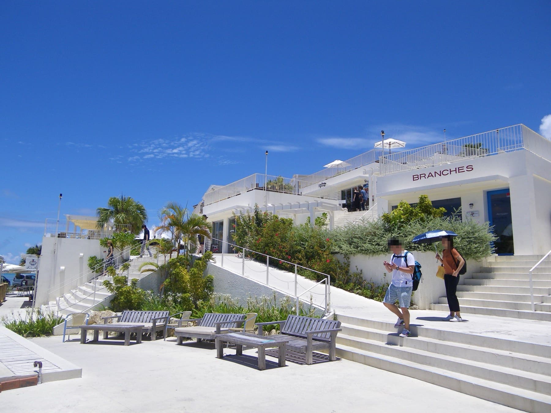 White terraced shops and cafes of Umikaji Terrace step up the slope of Senagajima under a deep blue sky with palms along the stairs
