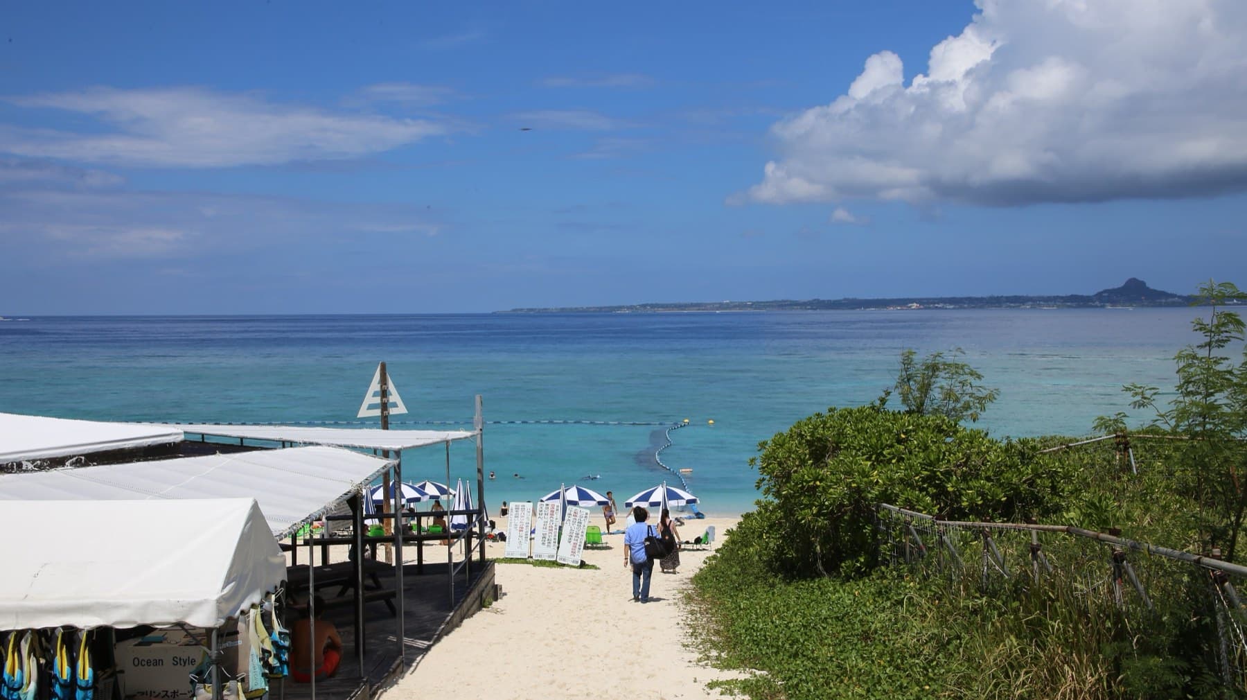 A sandy path leads past beach huts and parasols to Sesoko Beach, where the flat outline of Ie-jima and its single peak sit across the water