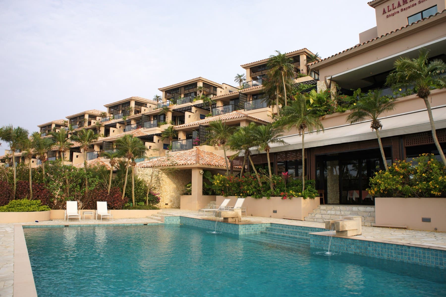 Sun loungers line a long blue pool below the stacked terraces and palms of Shigira Bayside Suite Allamanda.