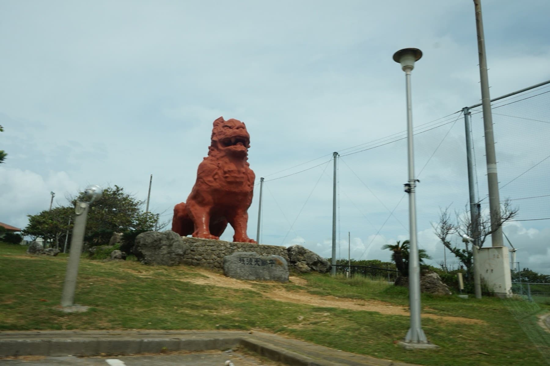 The giant red shisa statue of Zanpa Misaki Park sits on a rocky plinth above the grass on a cloudy day in Yomitan