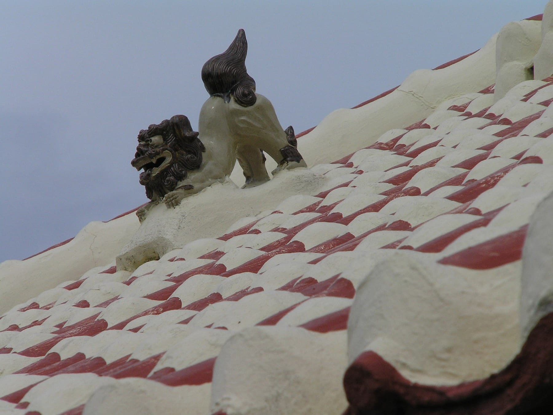 A grey clay shisa guards the ridge of a roof of red tiles set in thick white mortar on Kuroshima under a pale sky