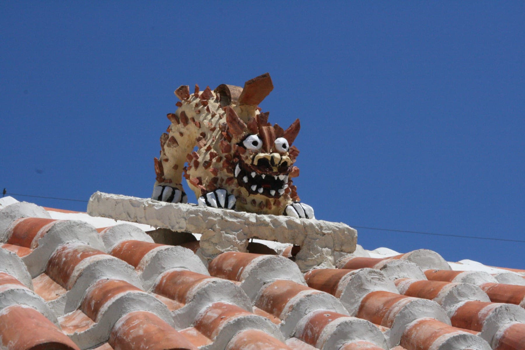 A fierce shisa made from shards of red tile and white mortar crouches on the ridge of a red-tile roof in Nago against a deep blue sky