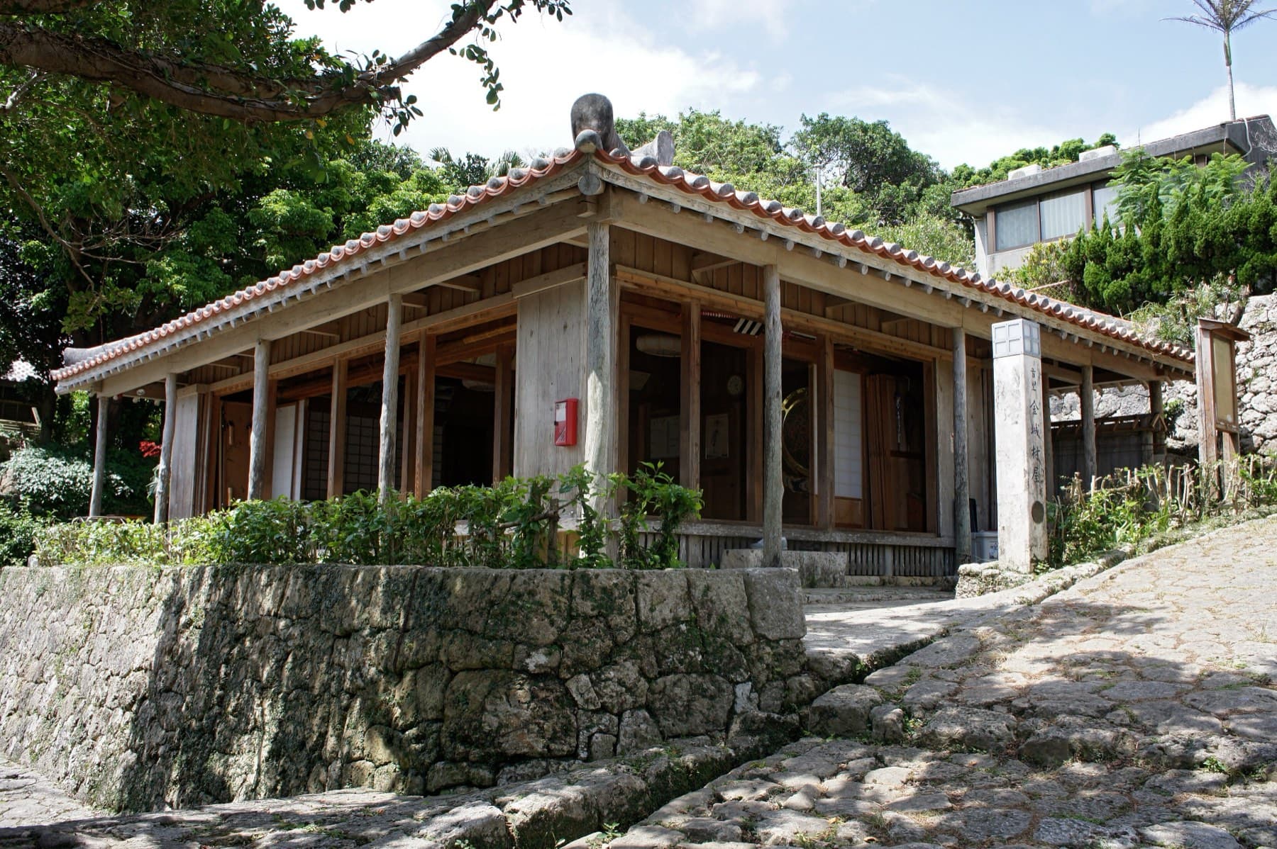 A wooden rest house with a red tile roof and open sides stands on a stone terrace beside the cobbles of the Kinjo-cho path