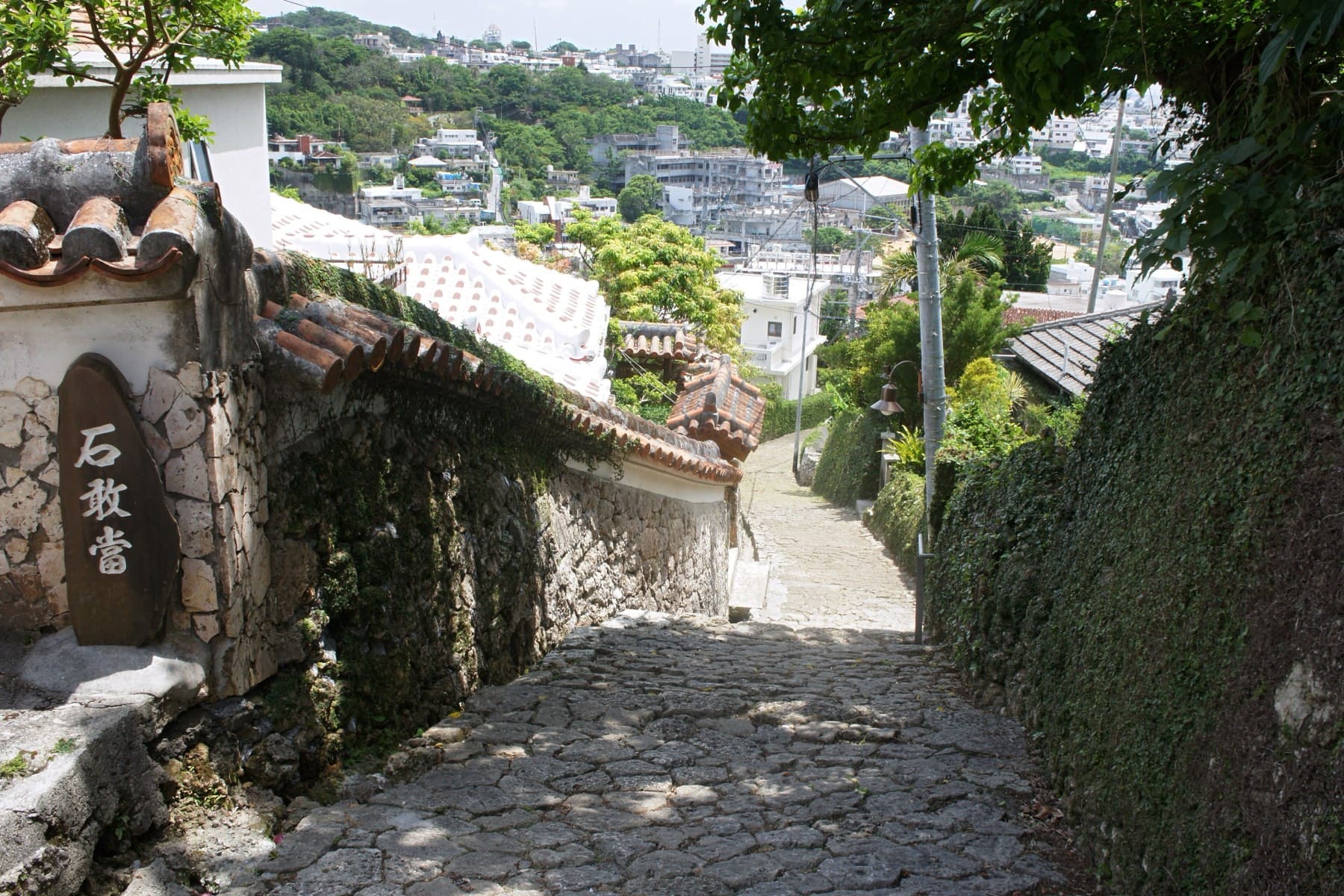 The Kinjo-cho stone path drops downhill between coral limestone walls and red tile roofs, and the hillside houses of Naha fill the view beyond