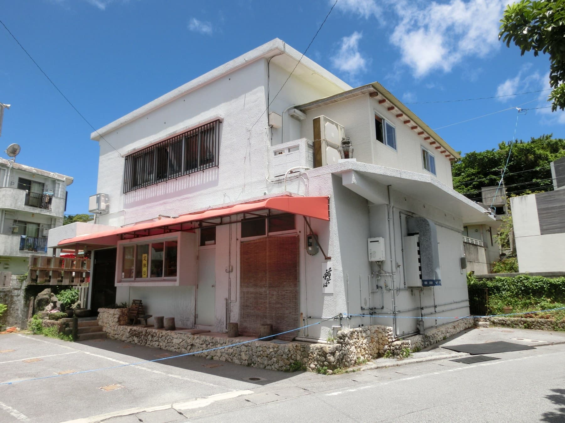 The white two-storey building that houses Shuri Soba sits on a quiet corner in Shuri Akata-cho under a blue sky, with a red awning over the shop front