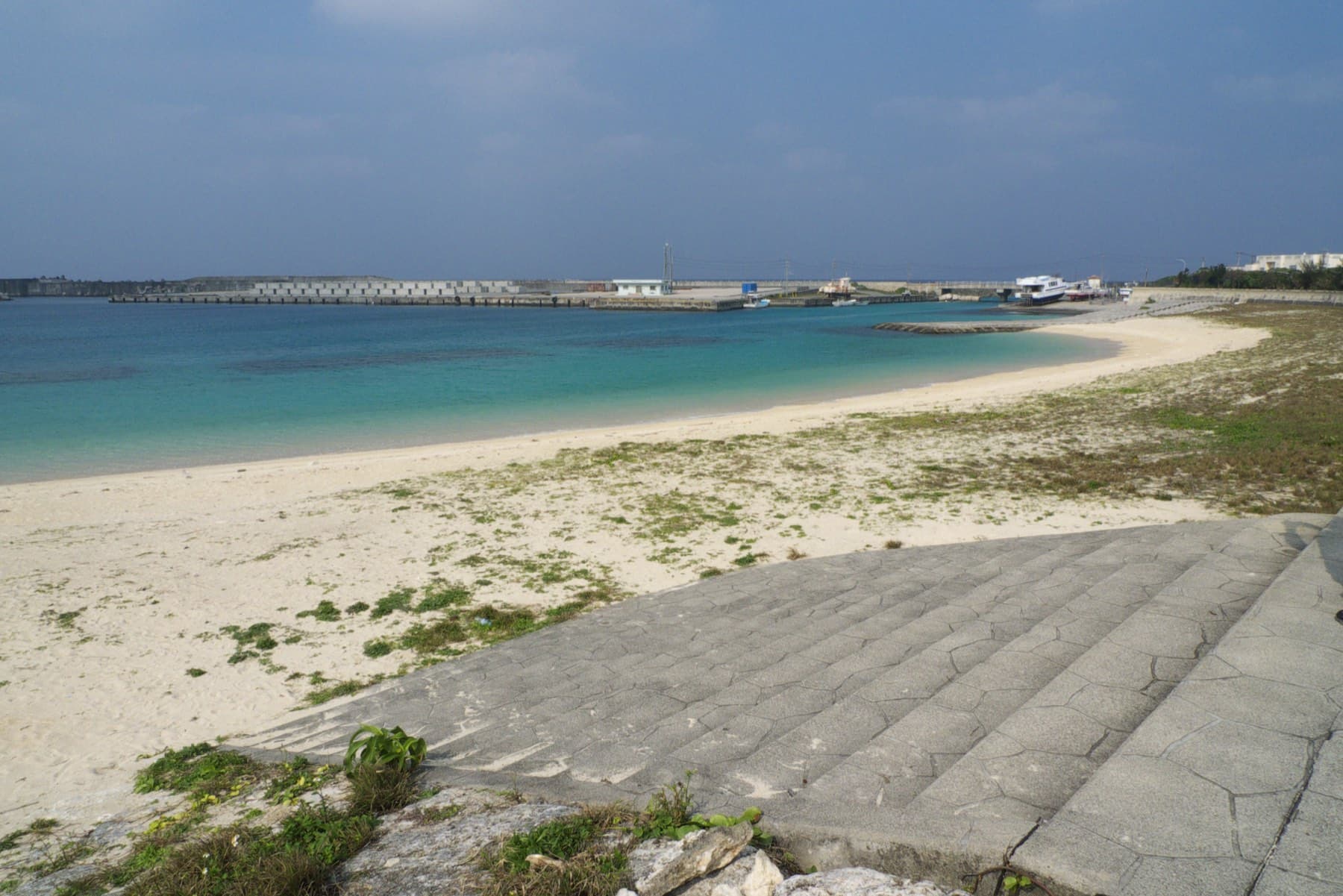 Nanta Beach curves from the seawall toward the breakwaters of Sonai port in bright, hazy light on the north coast of Yonaguni.