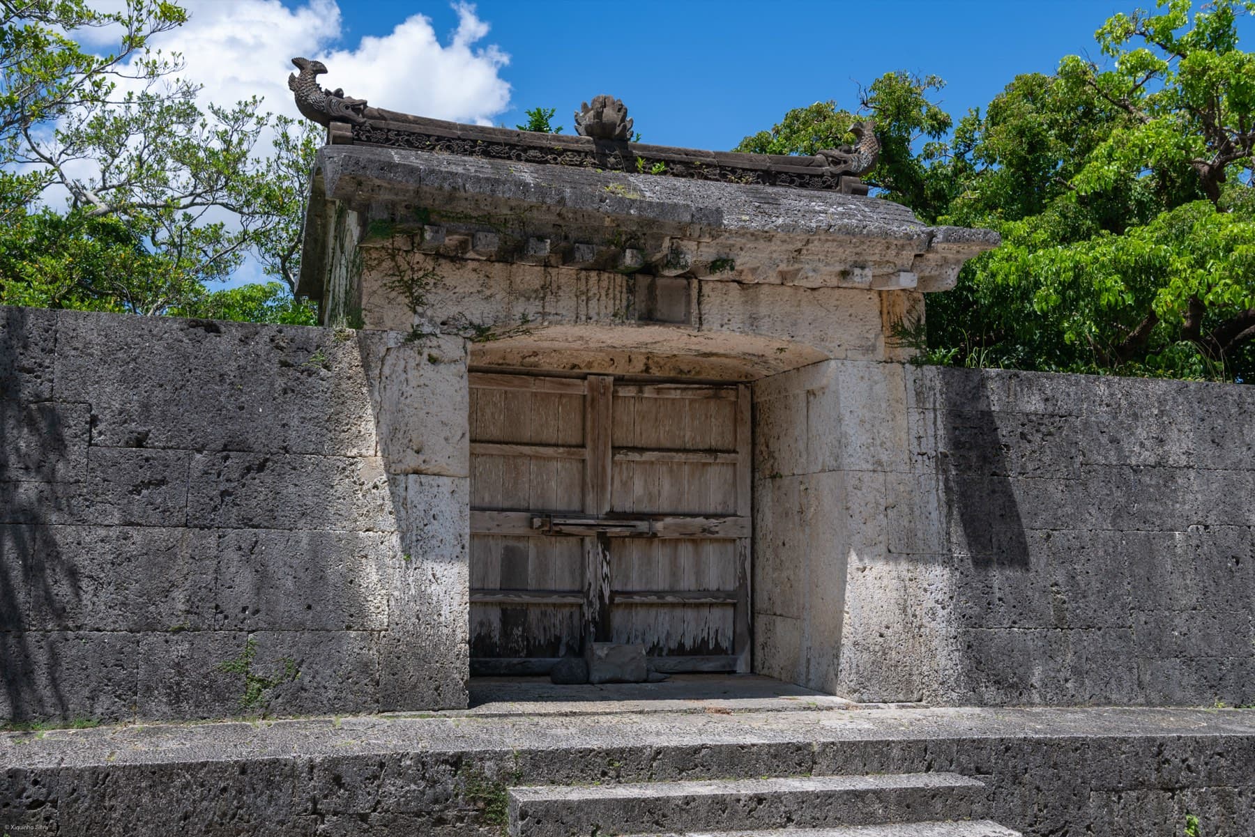 Carved coral limestone and a stone roof frame the weathered wooden doors of the Sonohyan-utaki gate on a bright summer day in Shuri