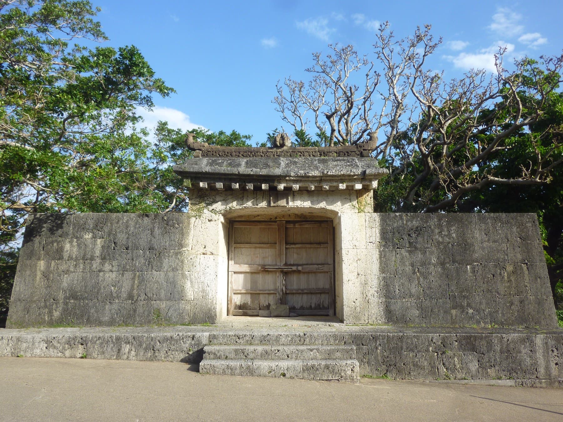 The closed stone gate of Sonohyan-utaki sits in a low limestone wall beside the path to Shuri Castle, and bare trees rise behind it under a blue sky