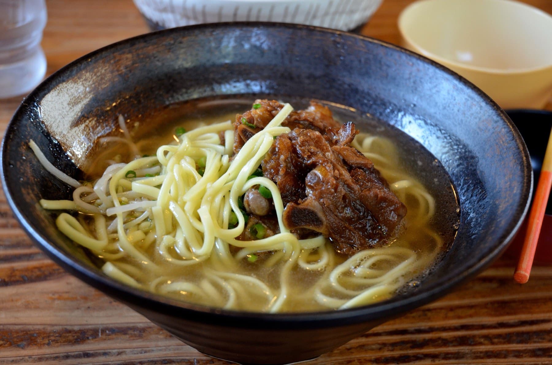 Stewed pork spare ribs and round yellow noodles fill a dark glazed bowl of clear broth on a wooden table at Takenoko on Taketomi