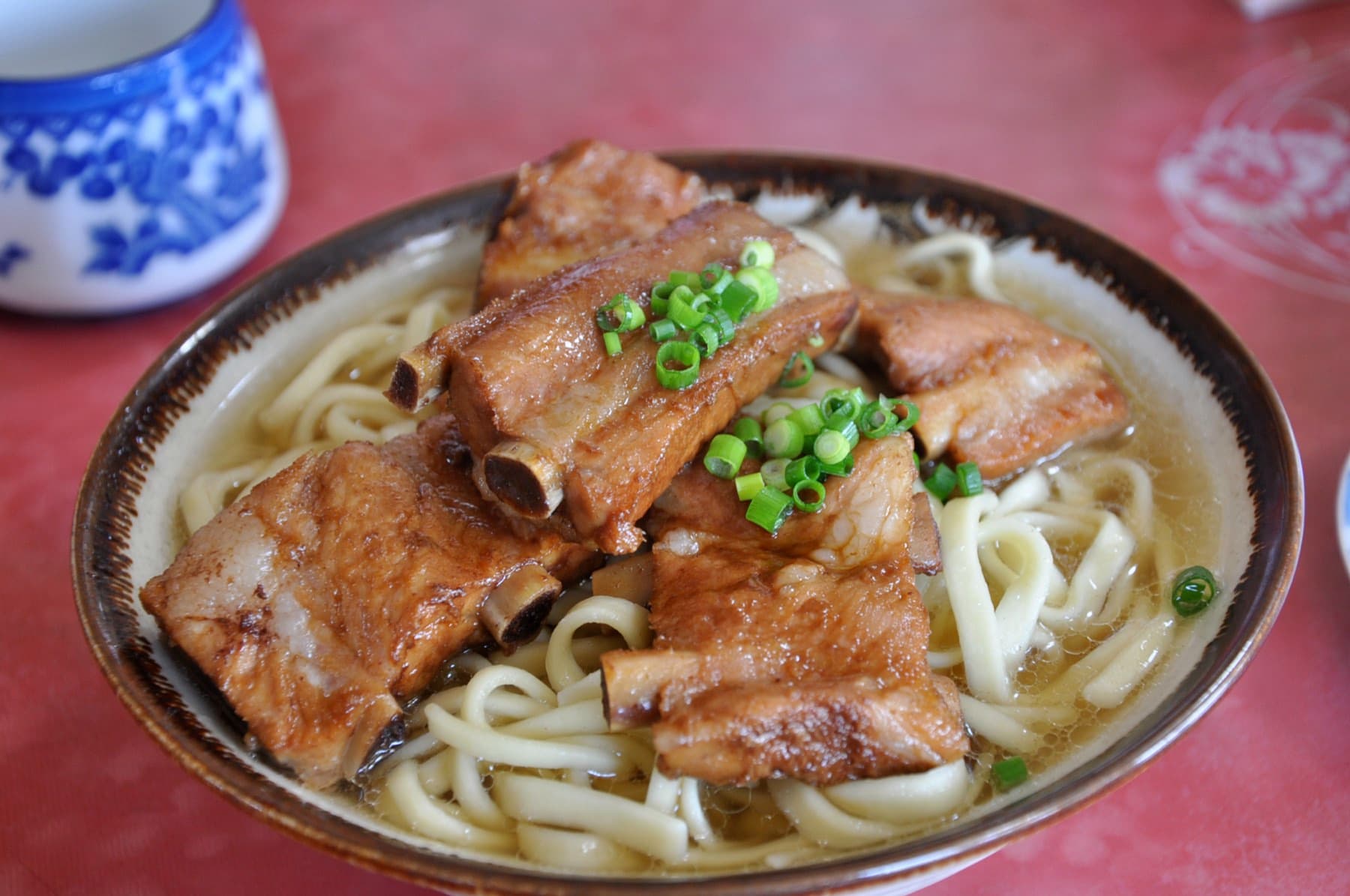 Glossy stewed pork spare ribs on the bone sit on thick wheat noodles with green onion in a bowl at Yanbaru Soba in Motobu