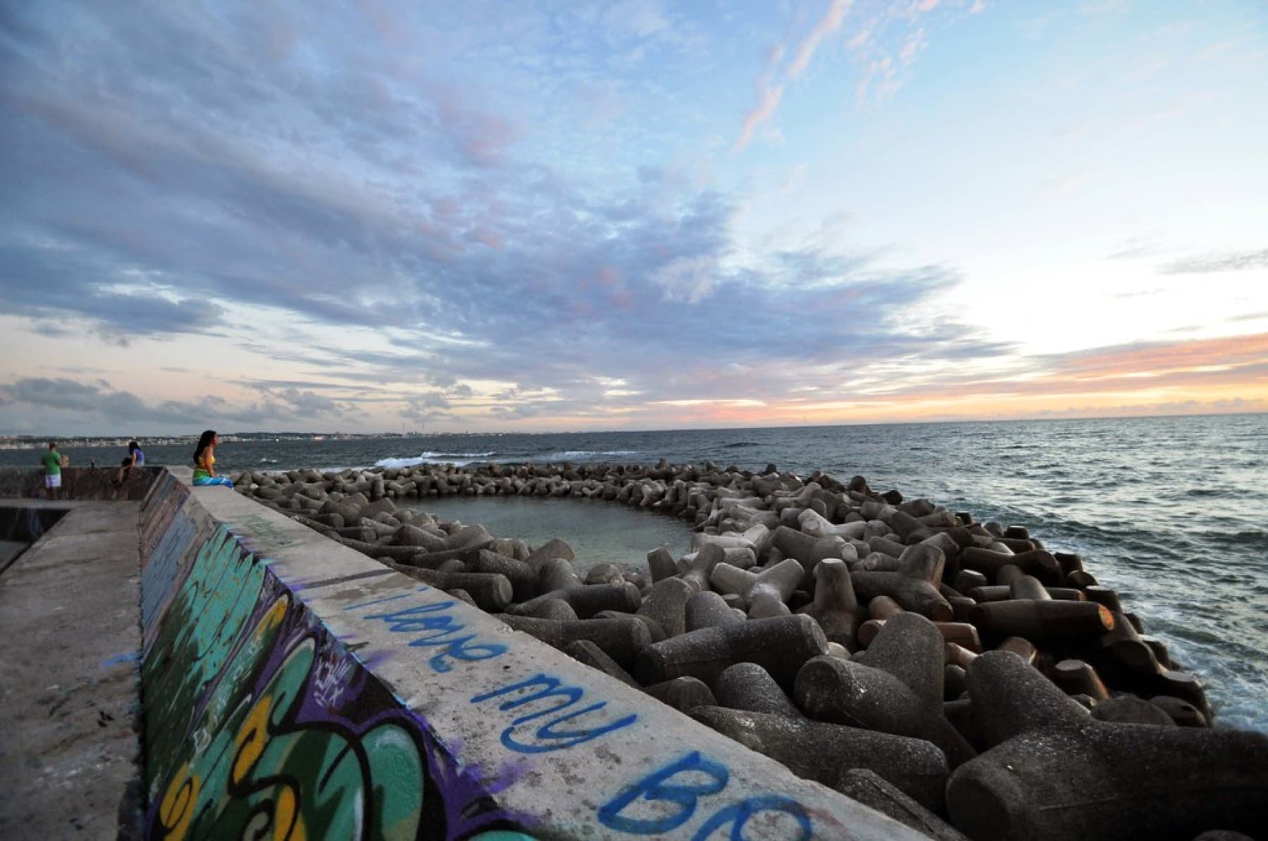 Painted graffiti covers the Sunabe seawall at dusk, and a curve of tetrapods holds back the waves under a soft pink and blue sky