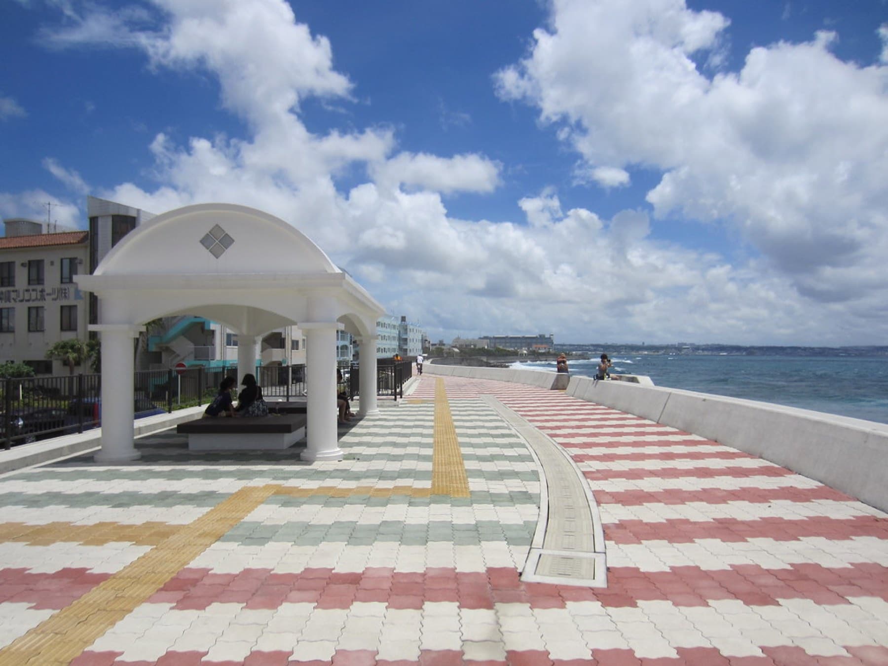 A white domed shelter stands on the tiled promenade of the Sunabe seawall, which runs along the open sea under bright summer cloud