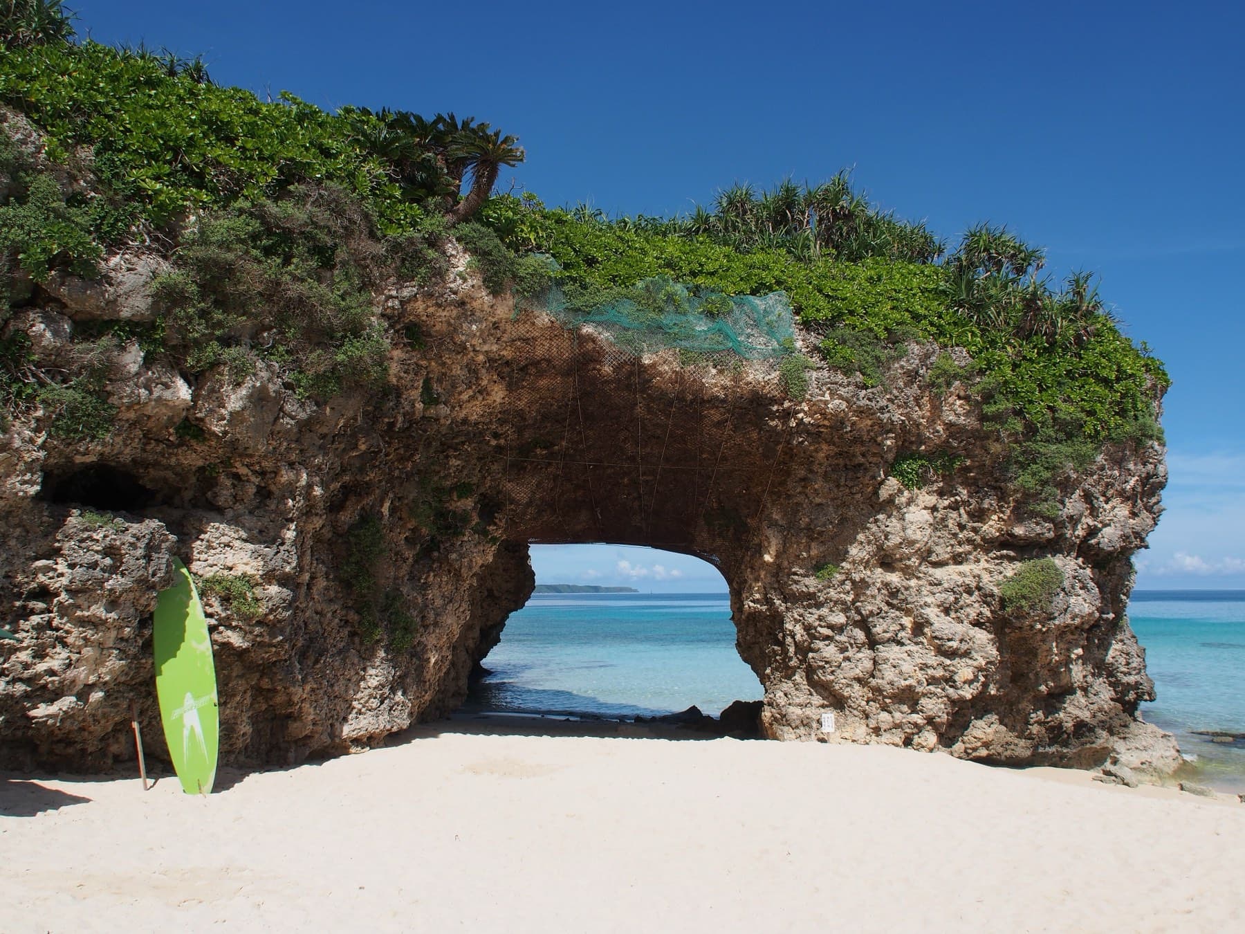 A great arch of weathered coral rock topped with green scrub frames the sea at Sunayama Beach under a clear blue sky.