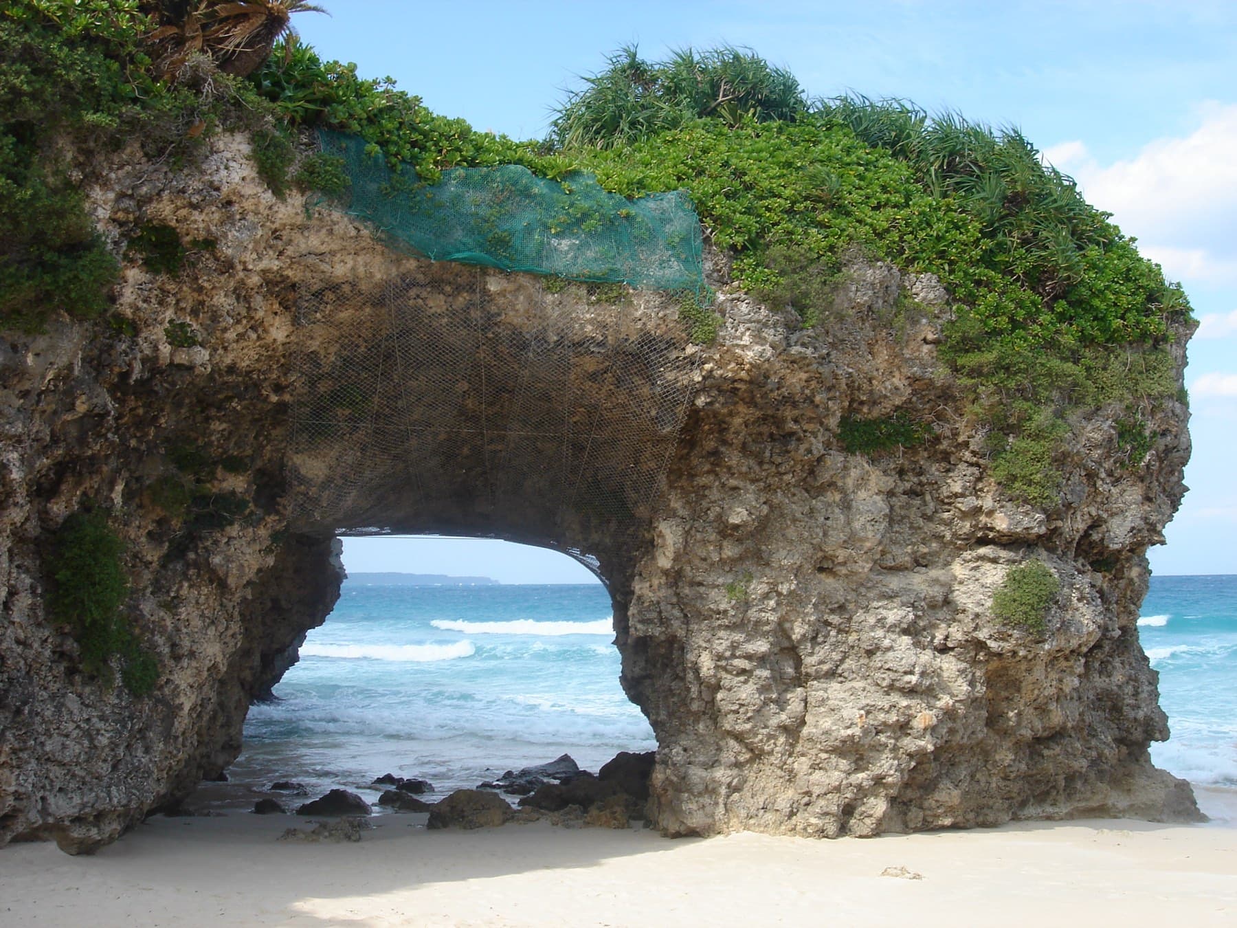 Small waves wash through the opening of the rock arch at Sunayama Beach, where the coral stone shows every pit and hollow.