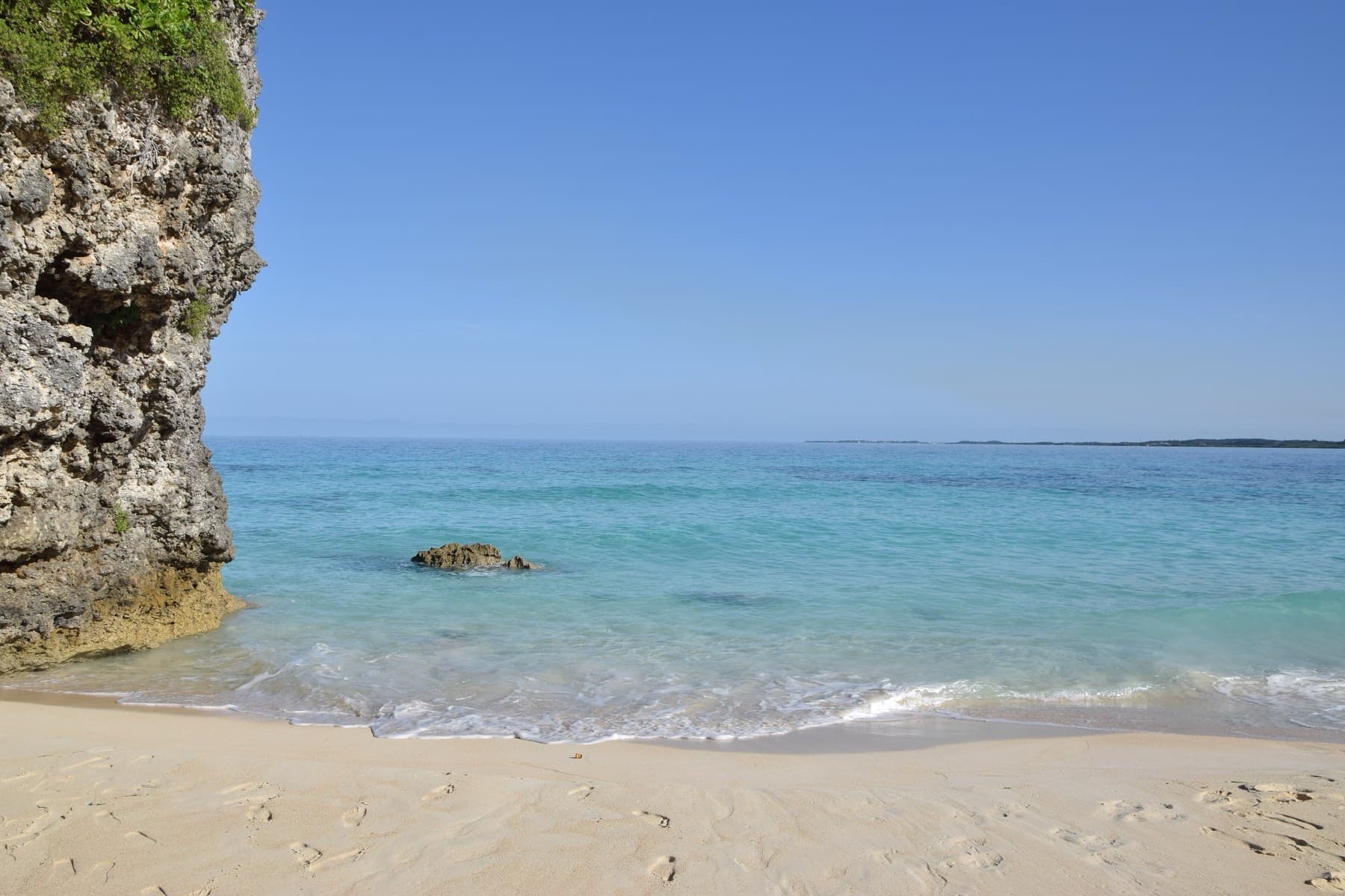 Clear water laps the pale sand beside a tall limestone wall at the end of Sunayama Beach on a still, cloudless day.