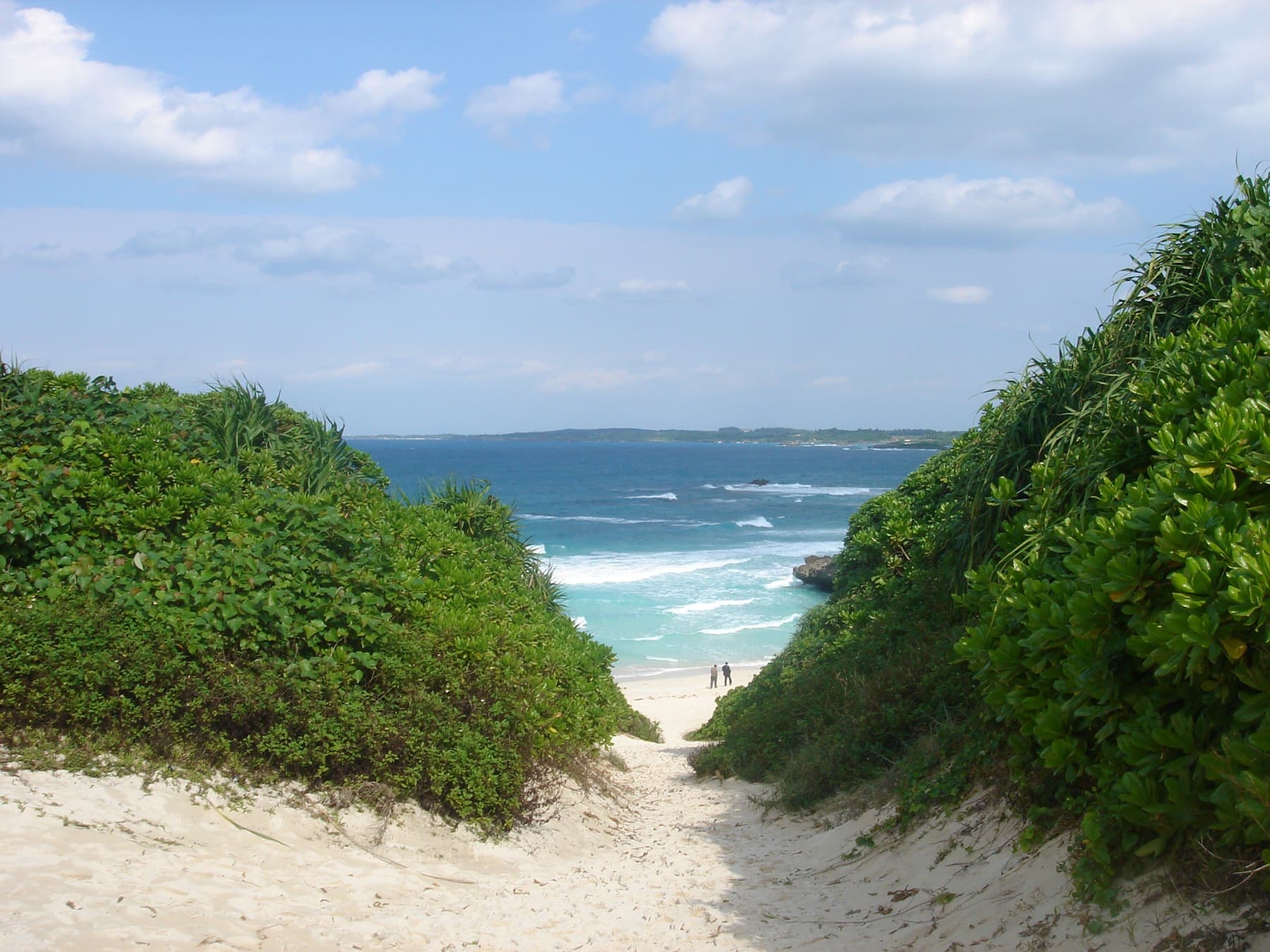 A sandy path drops between dune scrub to Sunayama Beach, where white surf rolls in over the reef and a few people stand at the water's edge.
