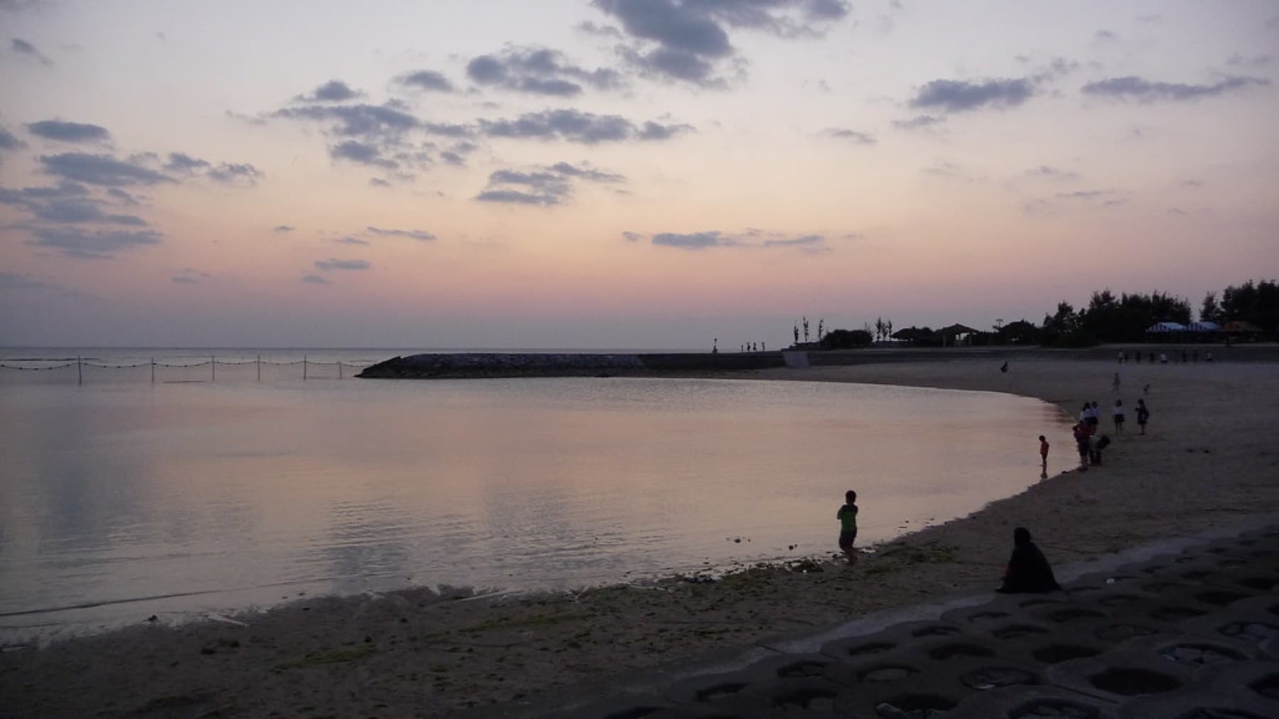 After sunset a few people wade at the edge of the calm curved bay of Chatan Sunset Beach under a pink and lilac sky