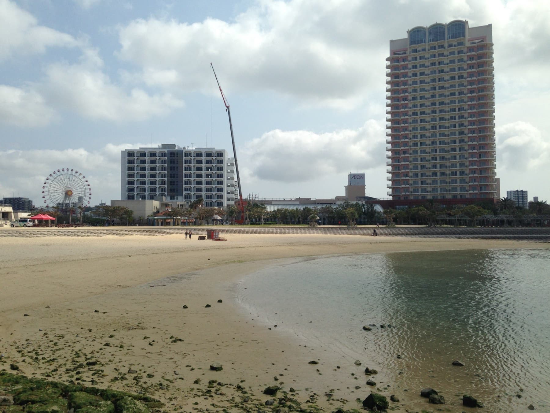 Low tide leaves wet sand and rock pools across Chatan Sunset Beach, with The Beach Tower Okinawa hotel and the American Village Ferris wheel behind