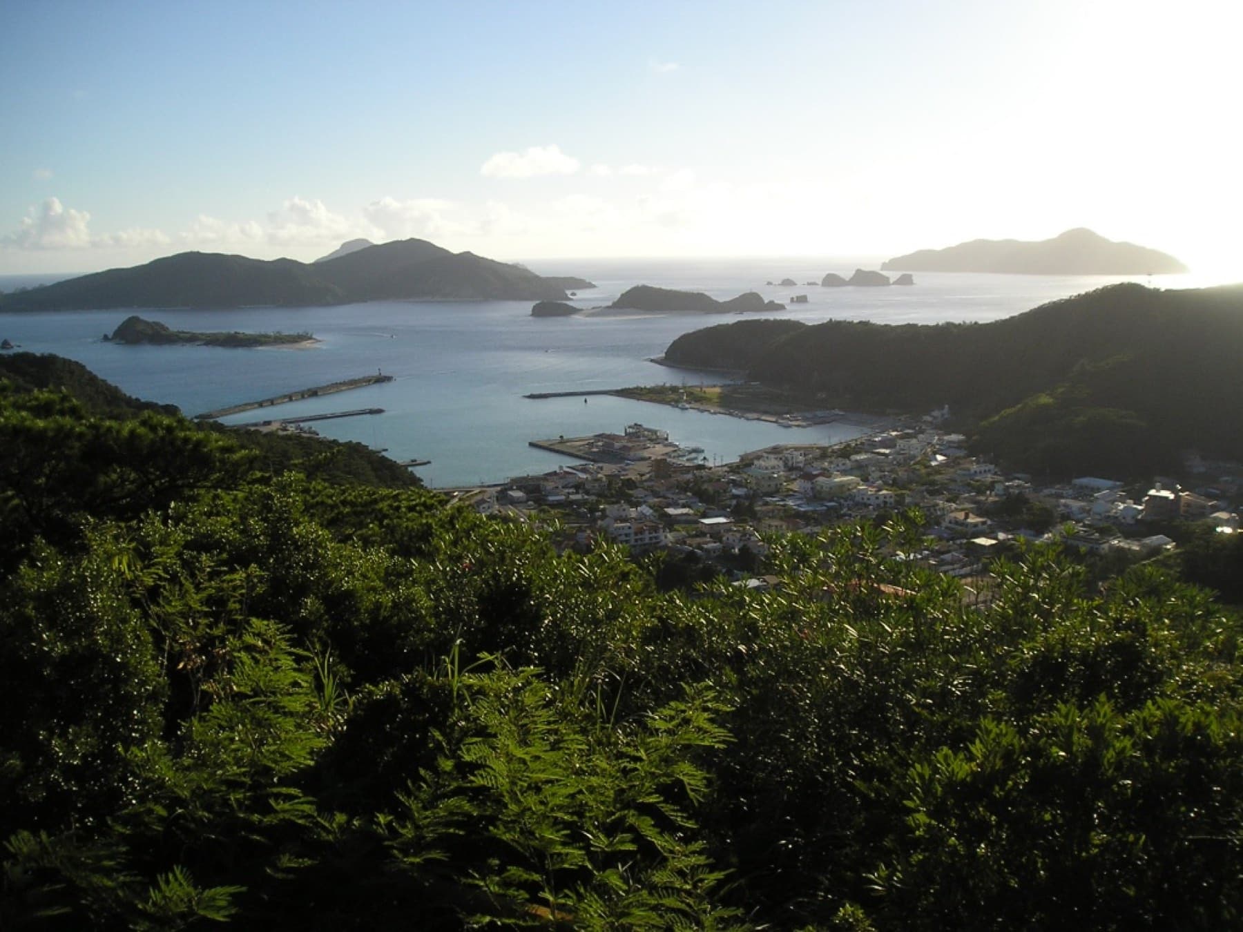 Late sun lights the harbour and rooftops of Zamami village below a forested hilltop, and Aka and smaller Kerama islets rise across the bay