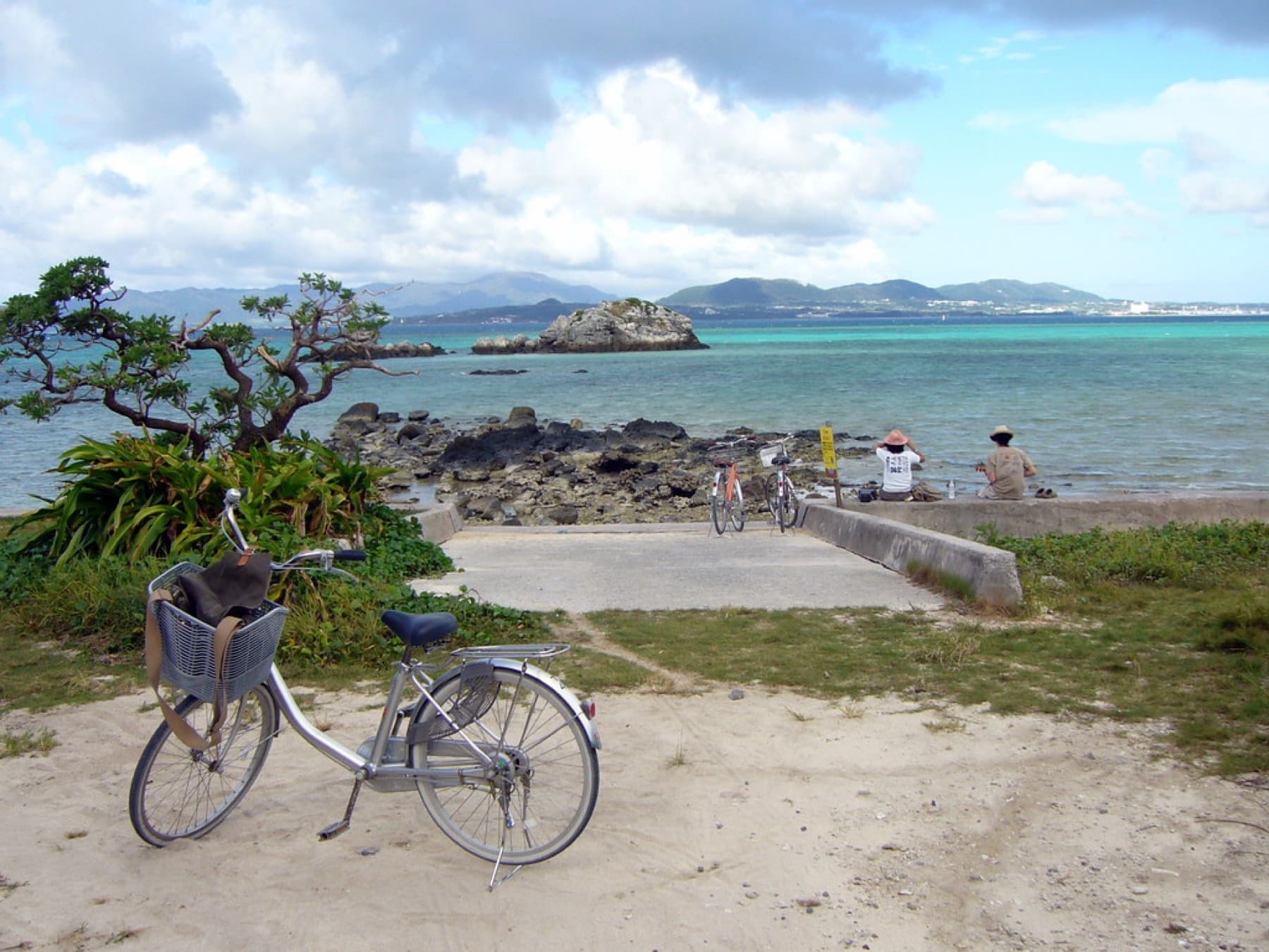 A rental bicycle stands on the sand above a slipway on Taketomi, and across the water lie a rocky islet and the long low shore of Ishigaki.