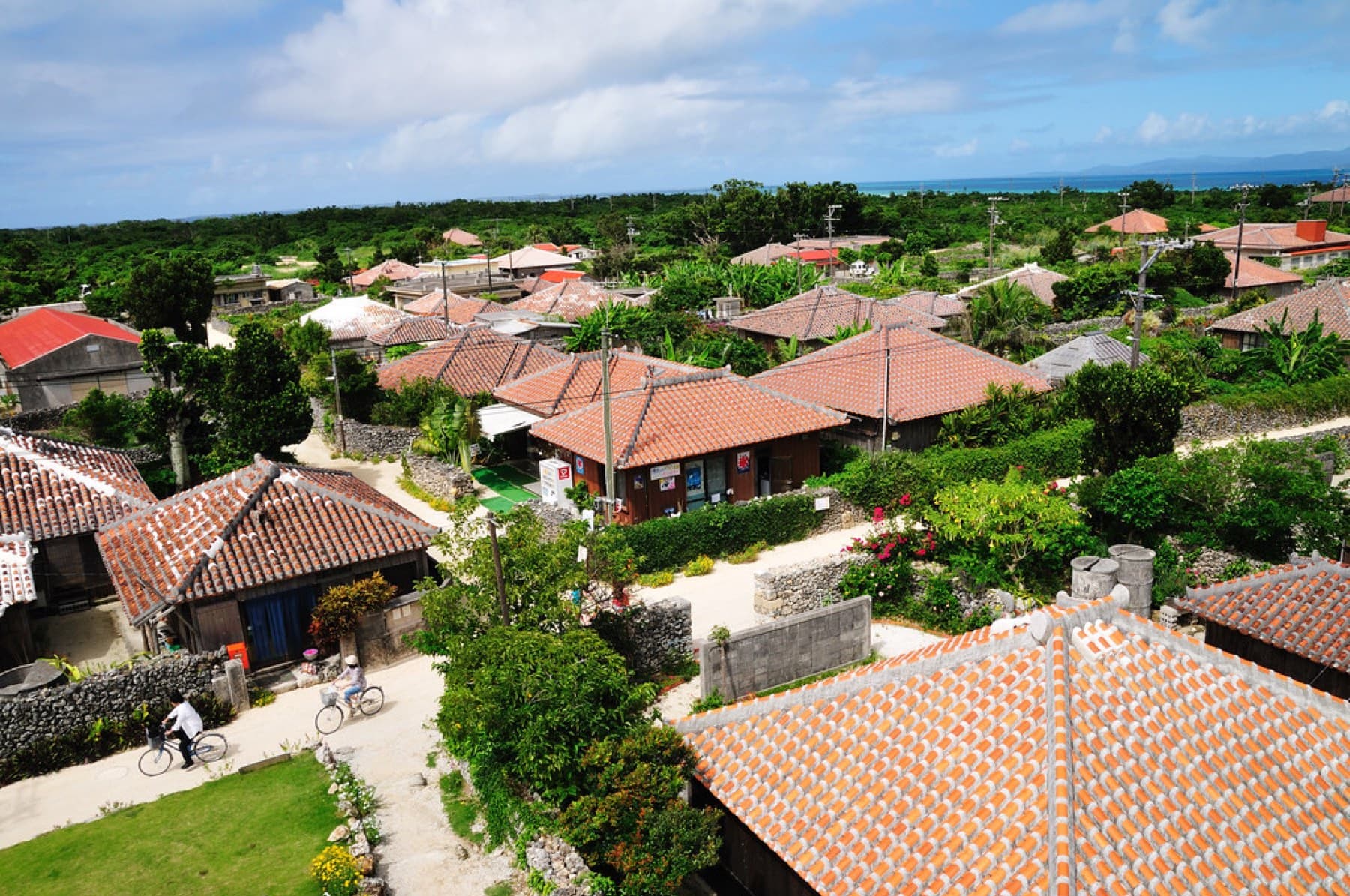 Red-tiled roofs and garden walls spread across Taketomi village under a summer sky while two cyclists roll along a white sand lane below.
