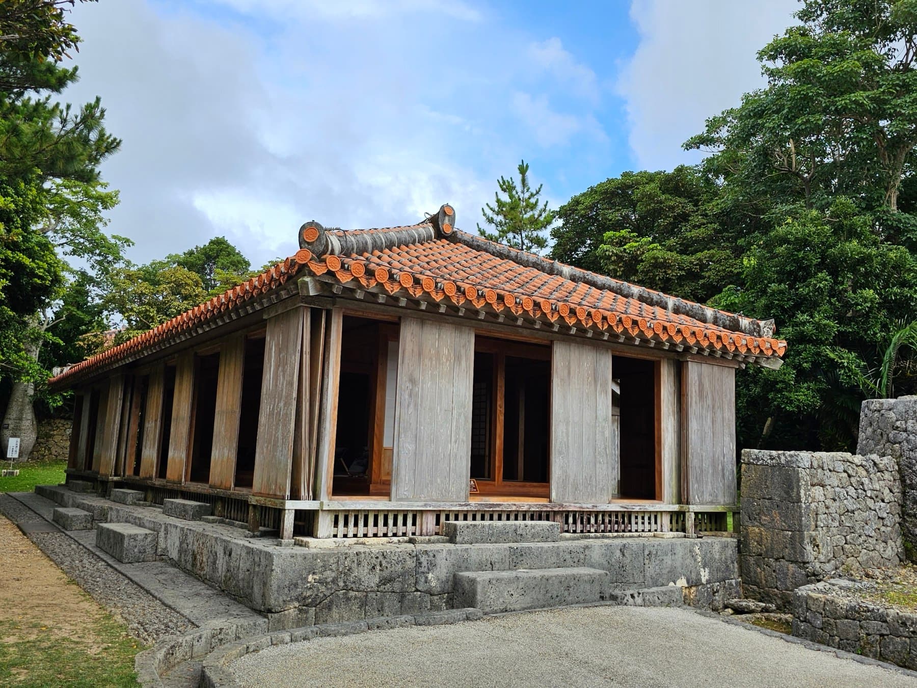 A wooden gatehouse with a red tile roof sits on a stone platform at the east side of the Tamaudun grounds, and tall trees shade it from behind
