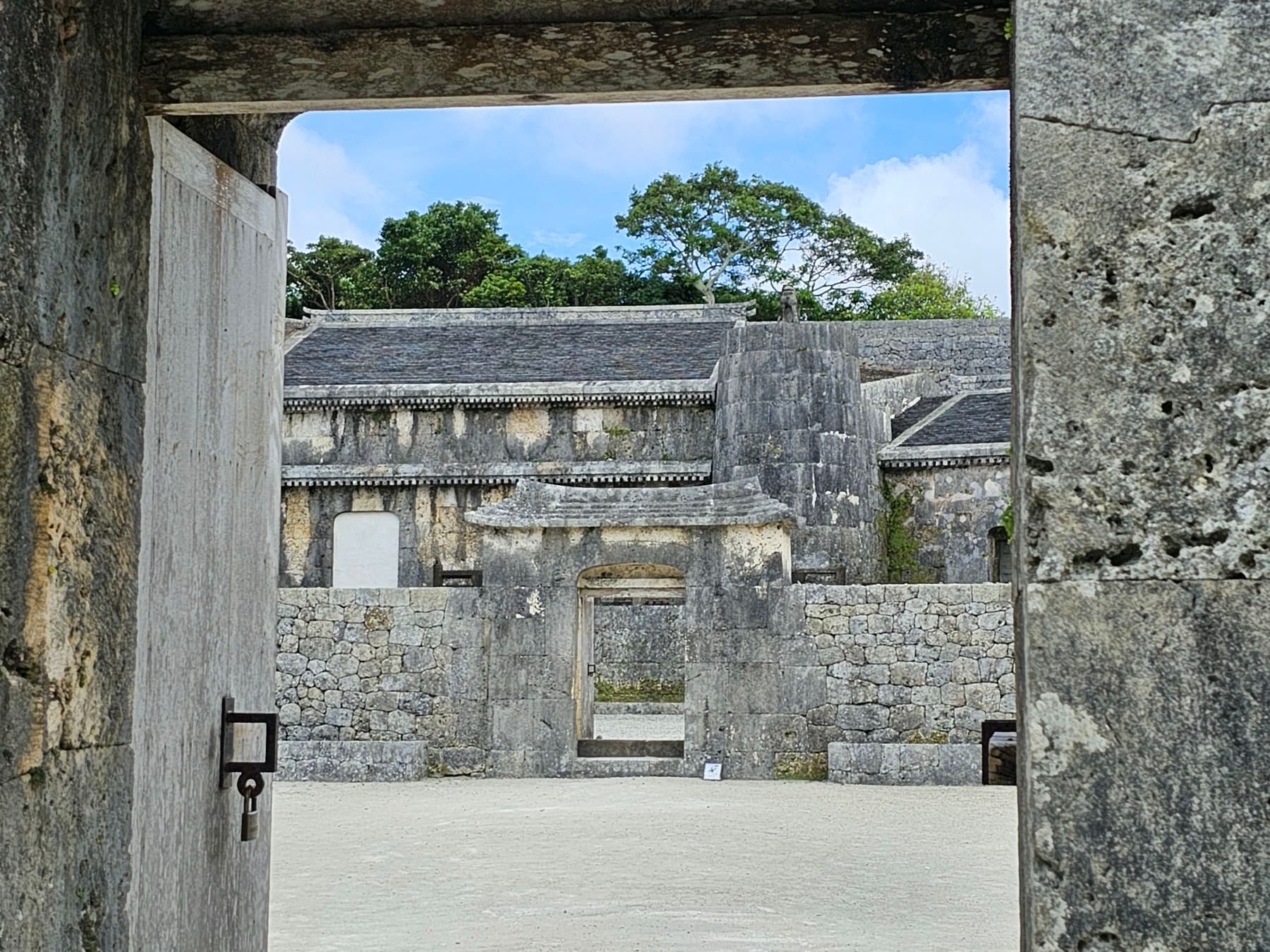 An open stone gateway at Tamaudun frames the inner wall and the left burial chamber of the royal tombs, and tall trees stand behind them