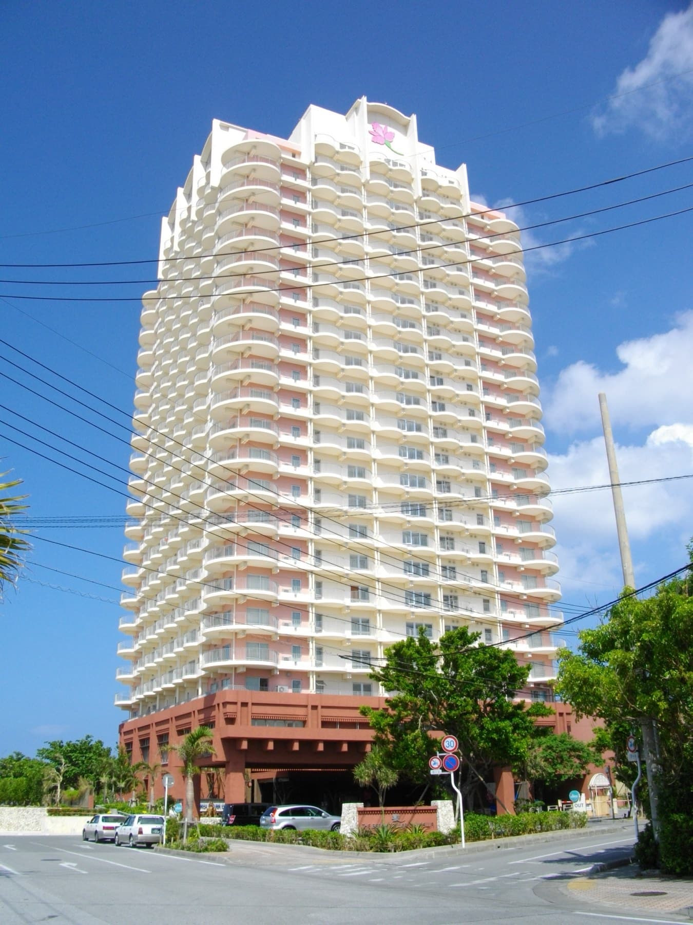 The tall cream tower of The Beach Tower Okinawa hotel rises above palm trees and the street in Mihama under a deep blue sky