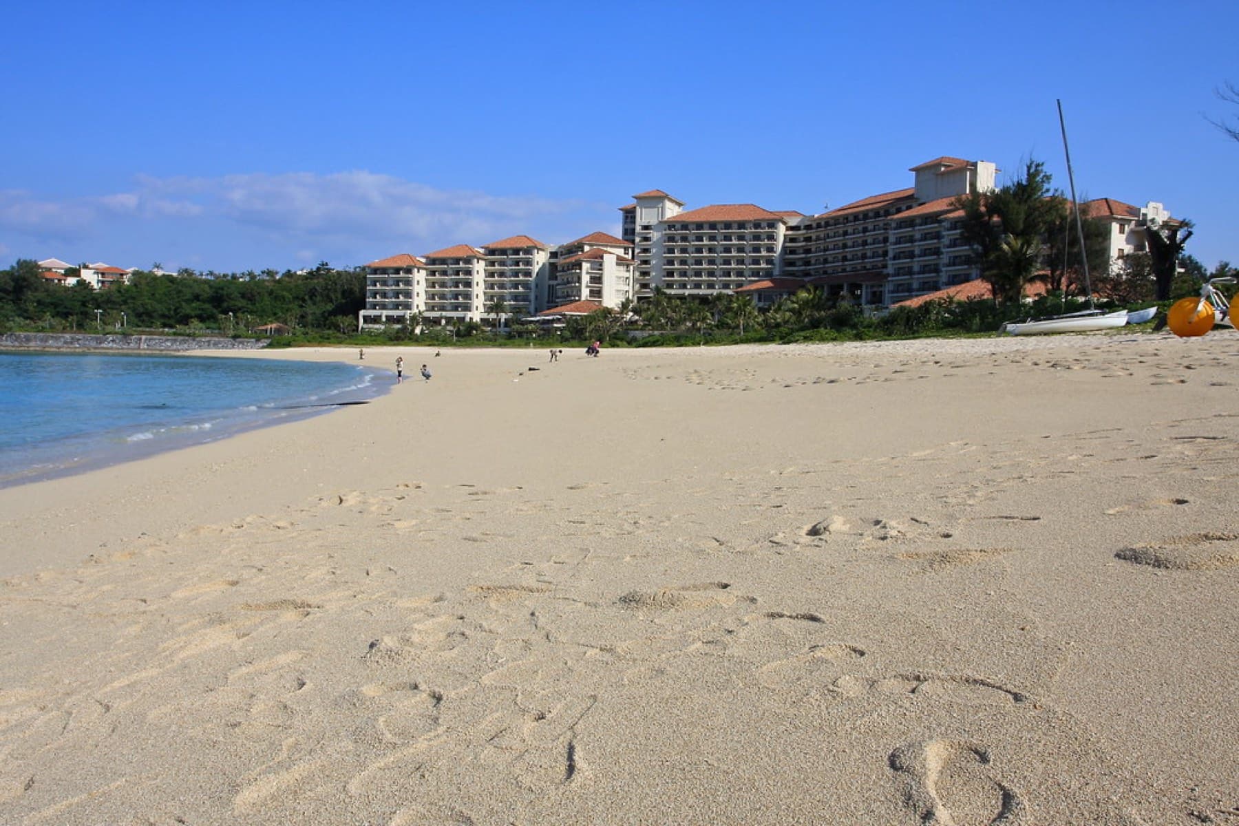 Footprints cross the wide pale beach in front of The Busena Terrace, whose red-roofed wings stand behind the sand on the Busena peninsula