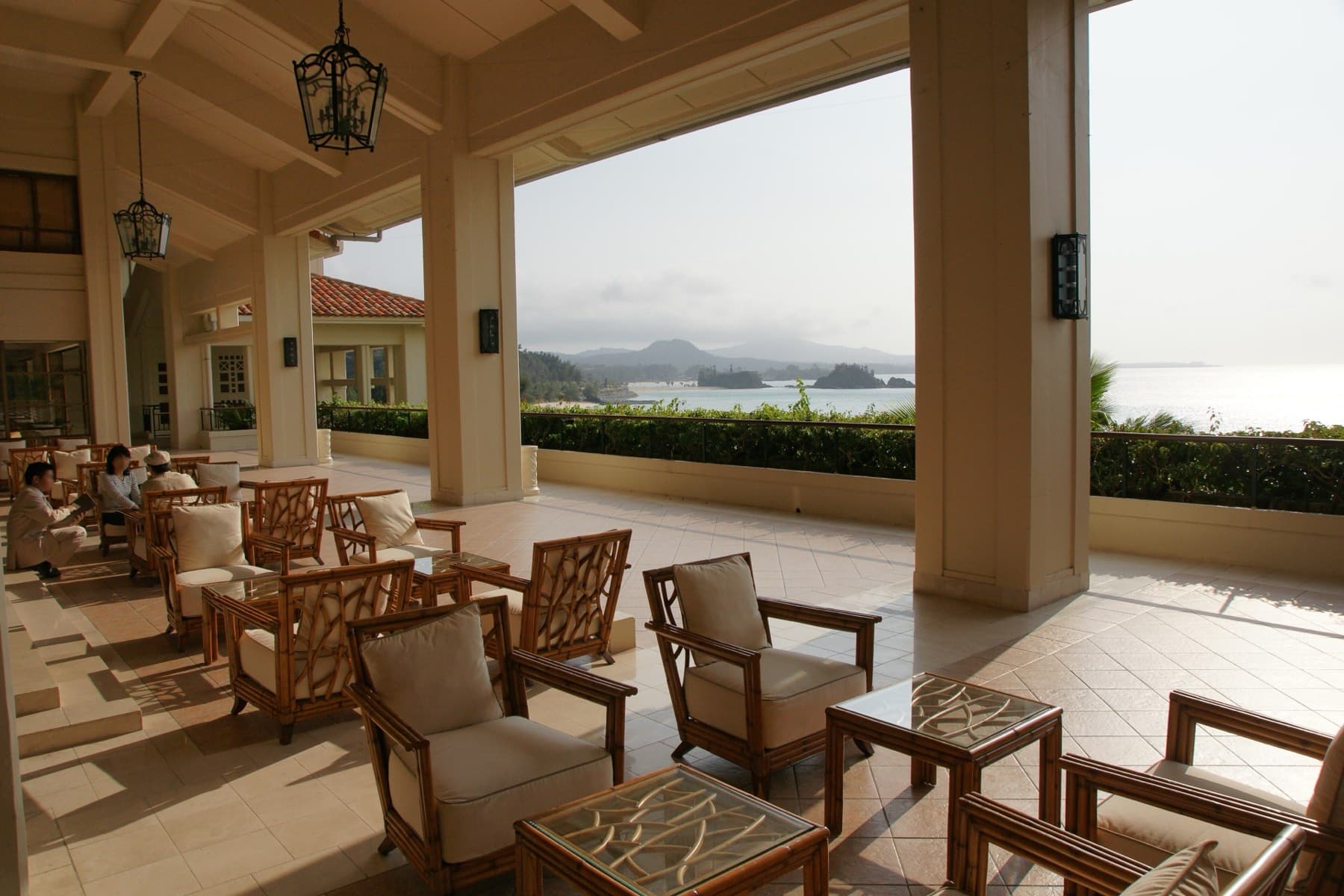 Wooden armchairs fill an open-sided lounge at The Busena Terrace, where tall columns frame a view of the bay and distant headlands