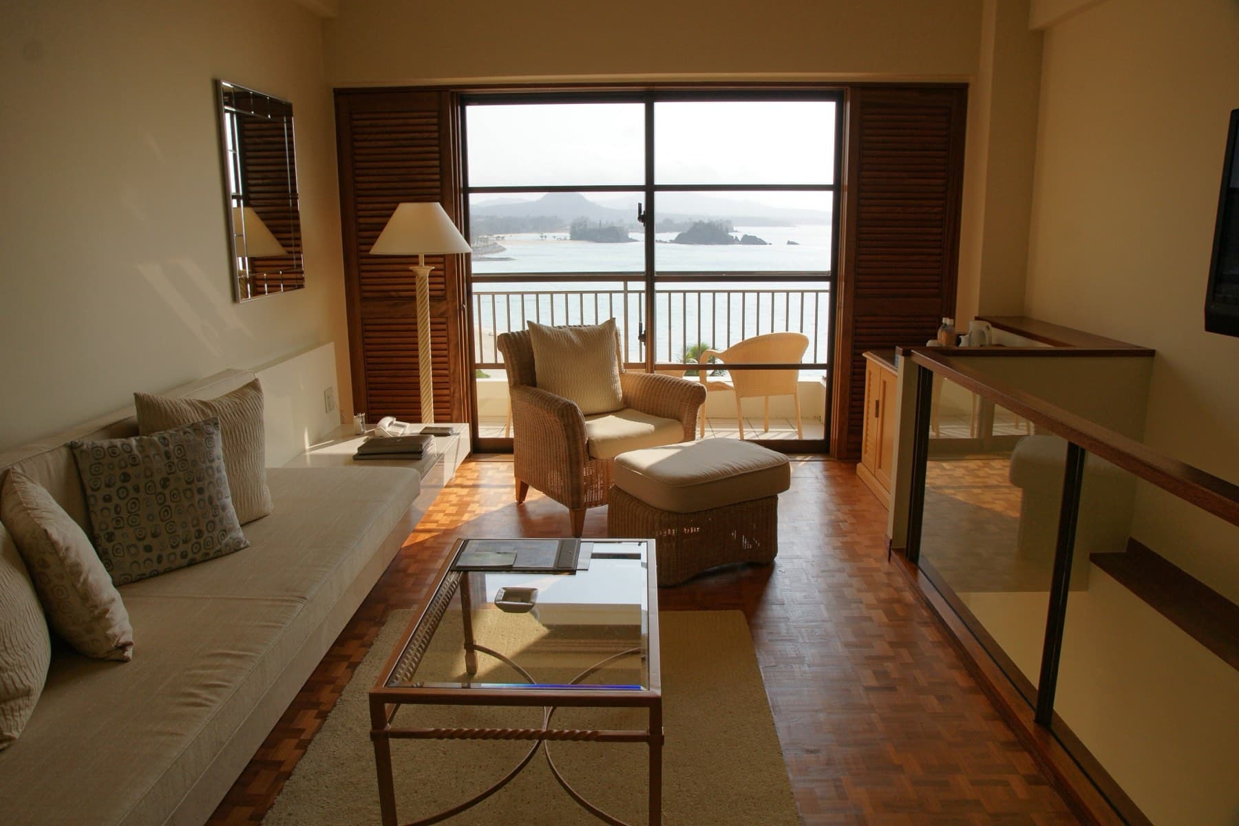A guest room at The Busena Terrace has pale sofas, a parquet floor and tall windows opening onto a balcony above wooded islets in the bay