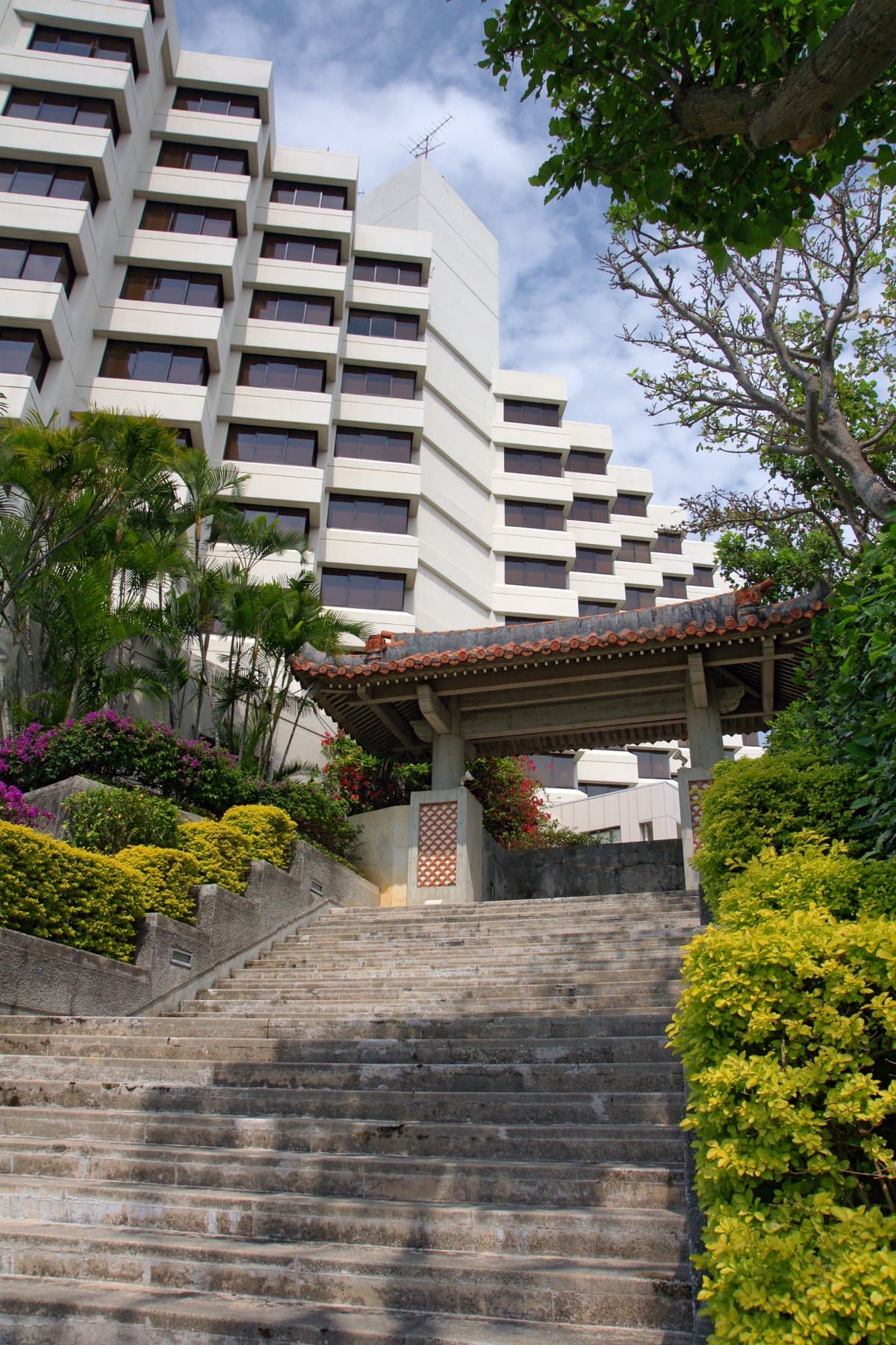 Stone steps climb between clipped hedges to a red-tiled gateway in front of the stepped white tower of The Naha Terrace hotel in Omoromachi
