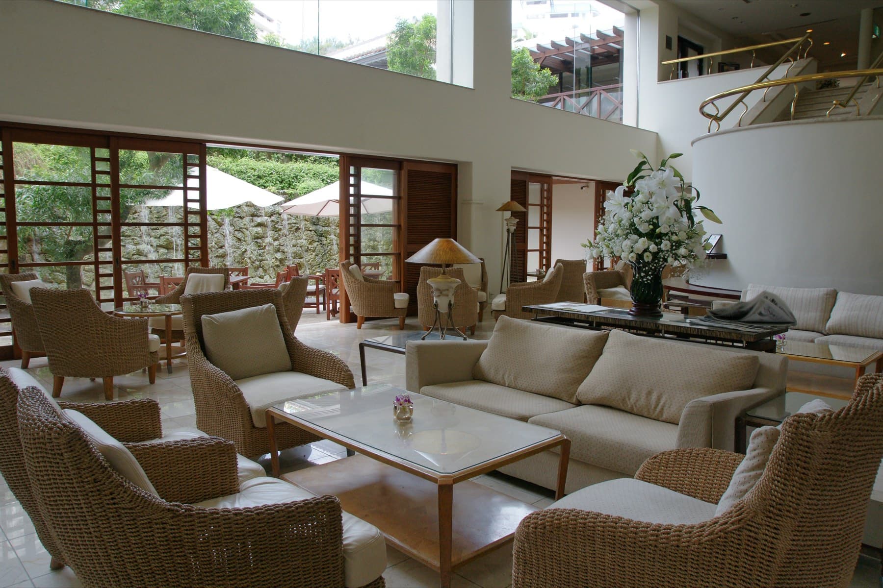 Rattan armchairs and cream sofas fill the bright lobby lounge of The Naha Terrace, where tall windows open onto a garden terrace with white umbrellas