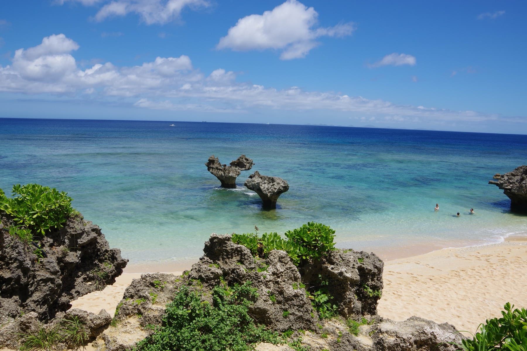 Dark coral rock and green scrub frame the sandy cove of Tinu Beach on Kouri, where Heart Rock stands in pale water and a few swimmers float nearby